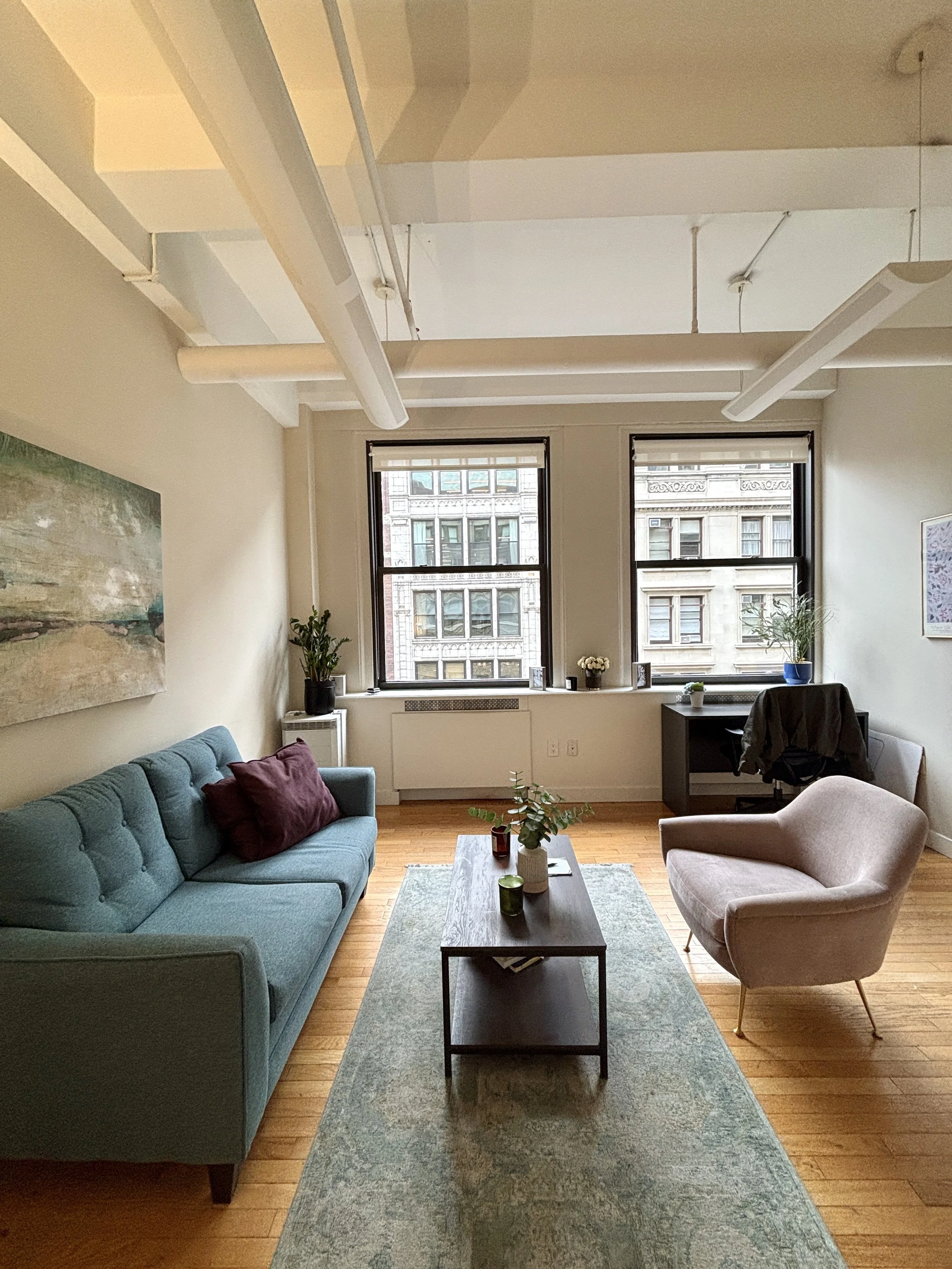 A living room with a green sofa, purple pillows, a beige armchair, a black coffee table with plants and candles, a vintage rug, large windows with city views, and a desk with a gray office chair.