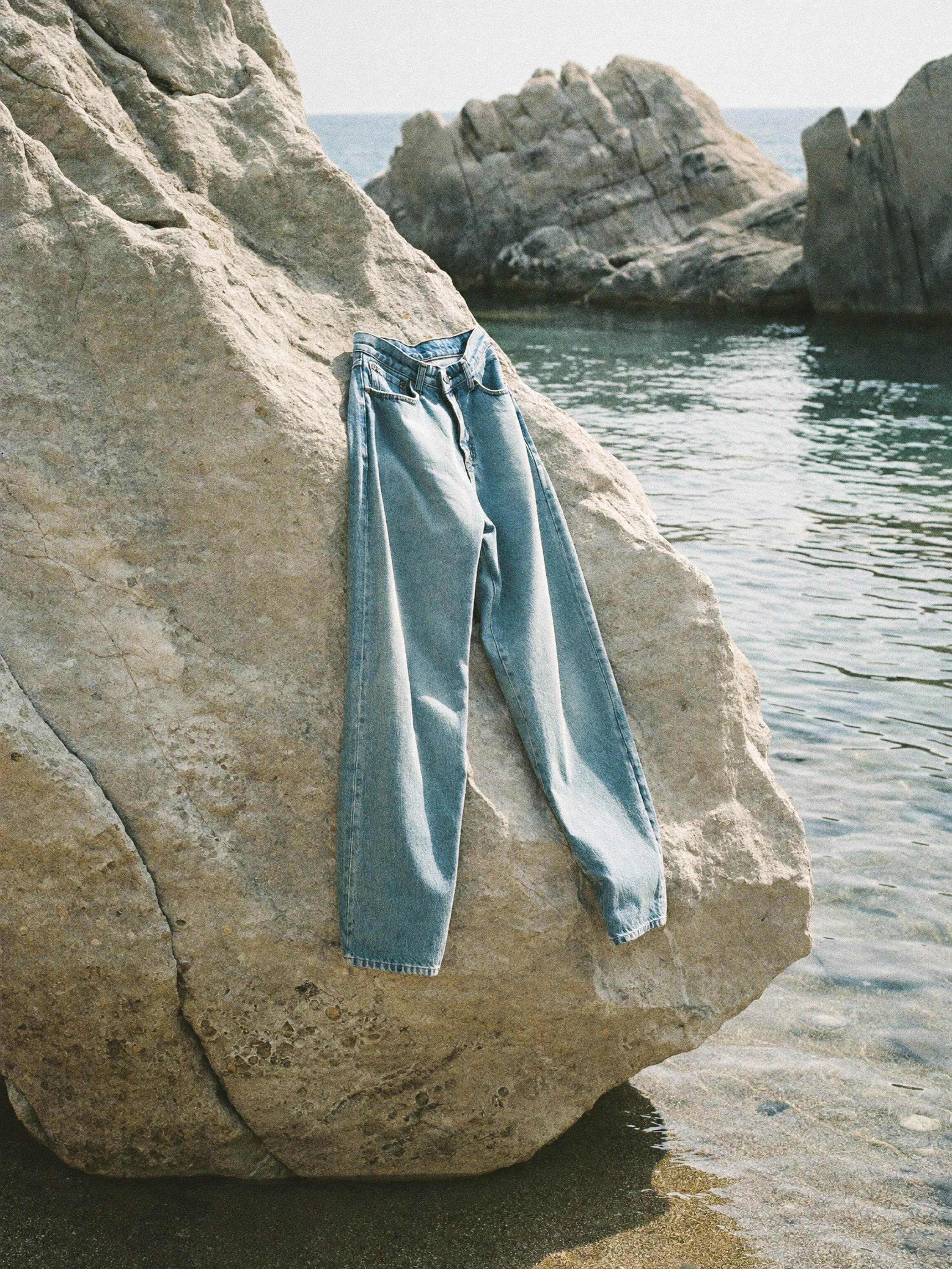 A pair of light blue jeans hanging over a large beige rock, with water and additional rocks in the background.
