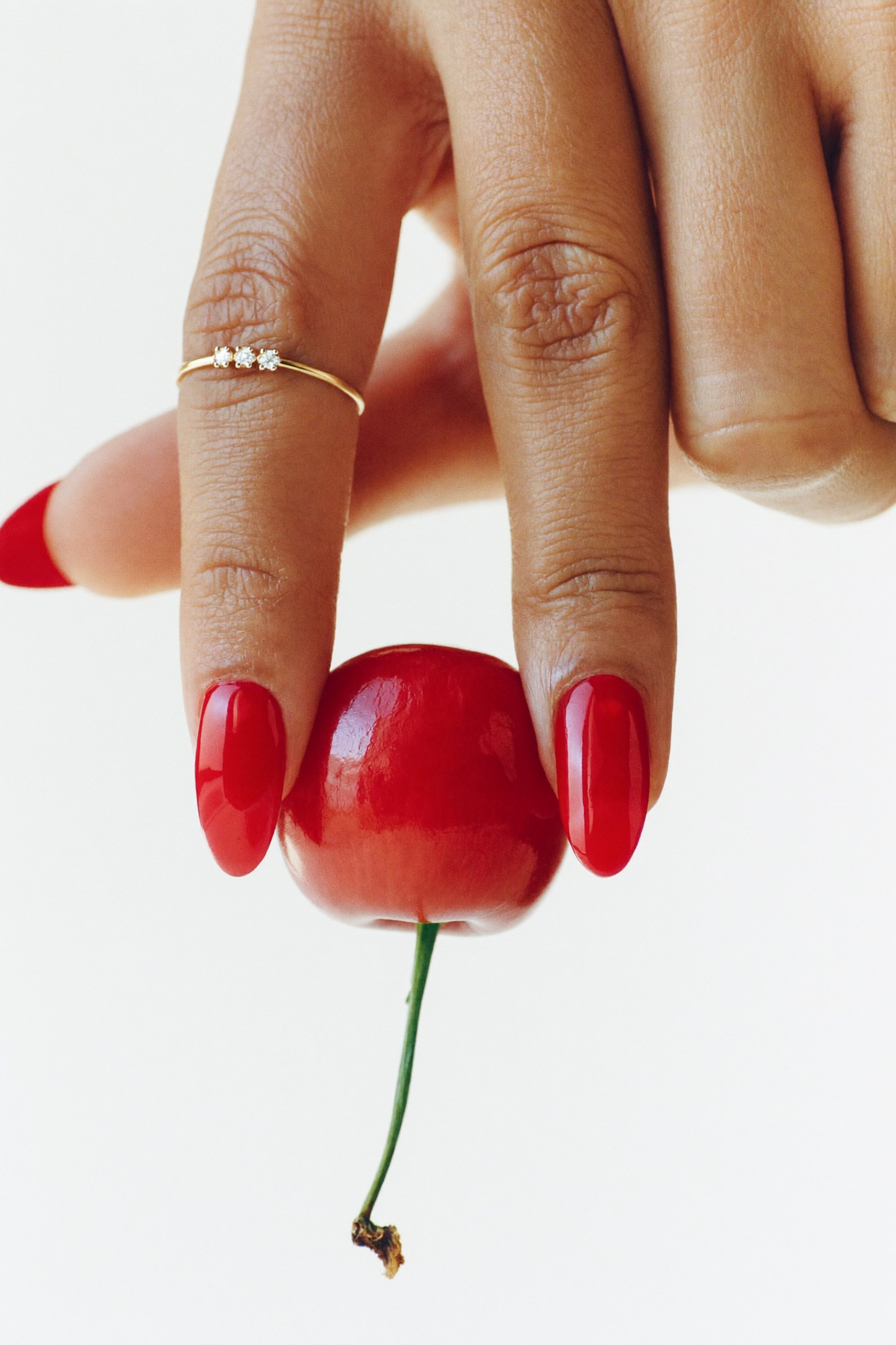 A person's hand with red-painted nails holding a cherry by its stem against a plain white background.
