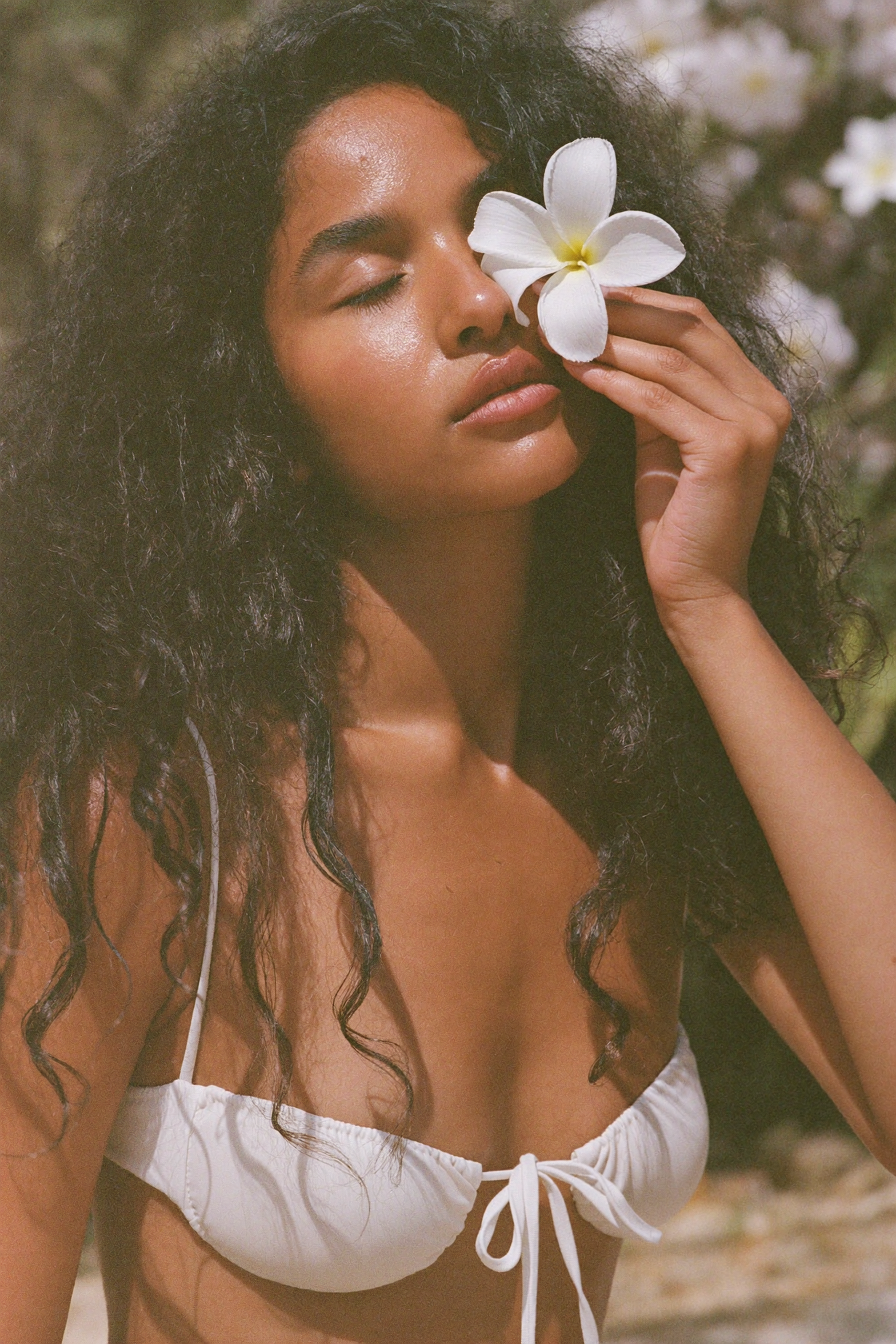A woman with curly hair and tan skin is outdoors, holding a white flower near her nose with her eyes closed, wearing a white bikini top with a bow.