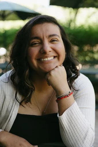 Smiling woman with dark wavy hair sitting outdoors with greenery in the background