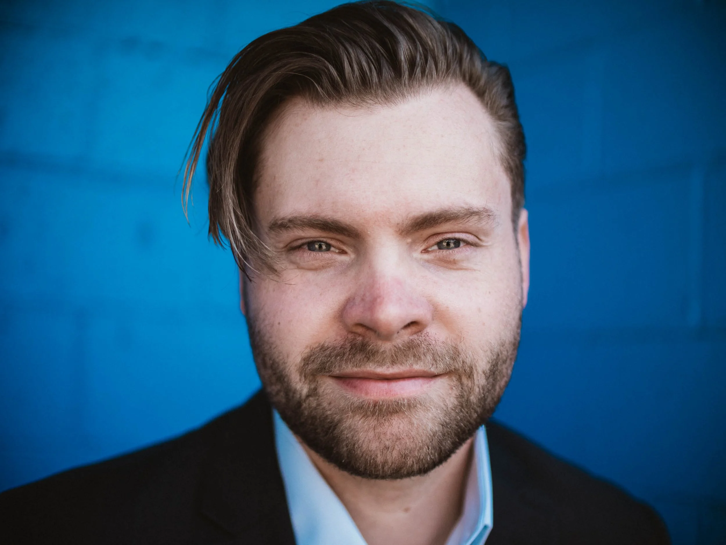 A close-up portrait of a man with brown hair styled to one side, wearing a black suit jacket and white shirt, smiling softly against a blue background.