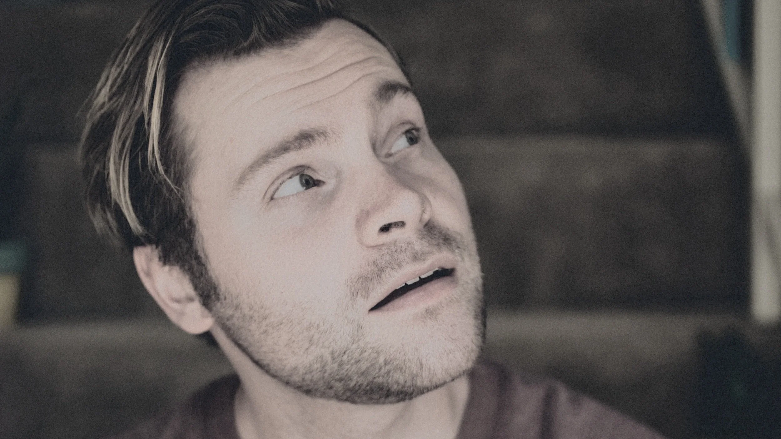 Close-up of a young man's face with light facial hair, looking upward and slightly to the side with a surprised or concerned expression.