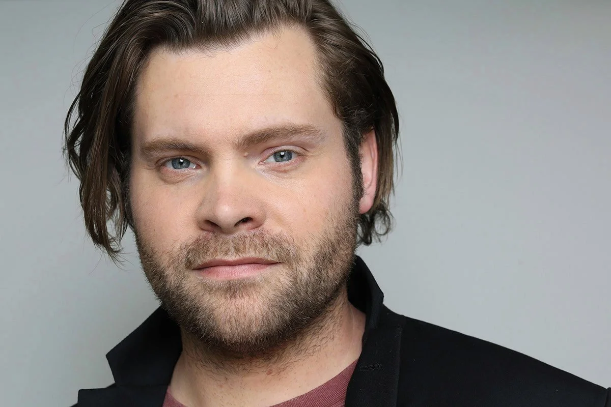 Close-up portrait of a man with medium-length brown hair, blue eyes, a beard, wearing a black jacket, against a gray background.