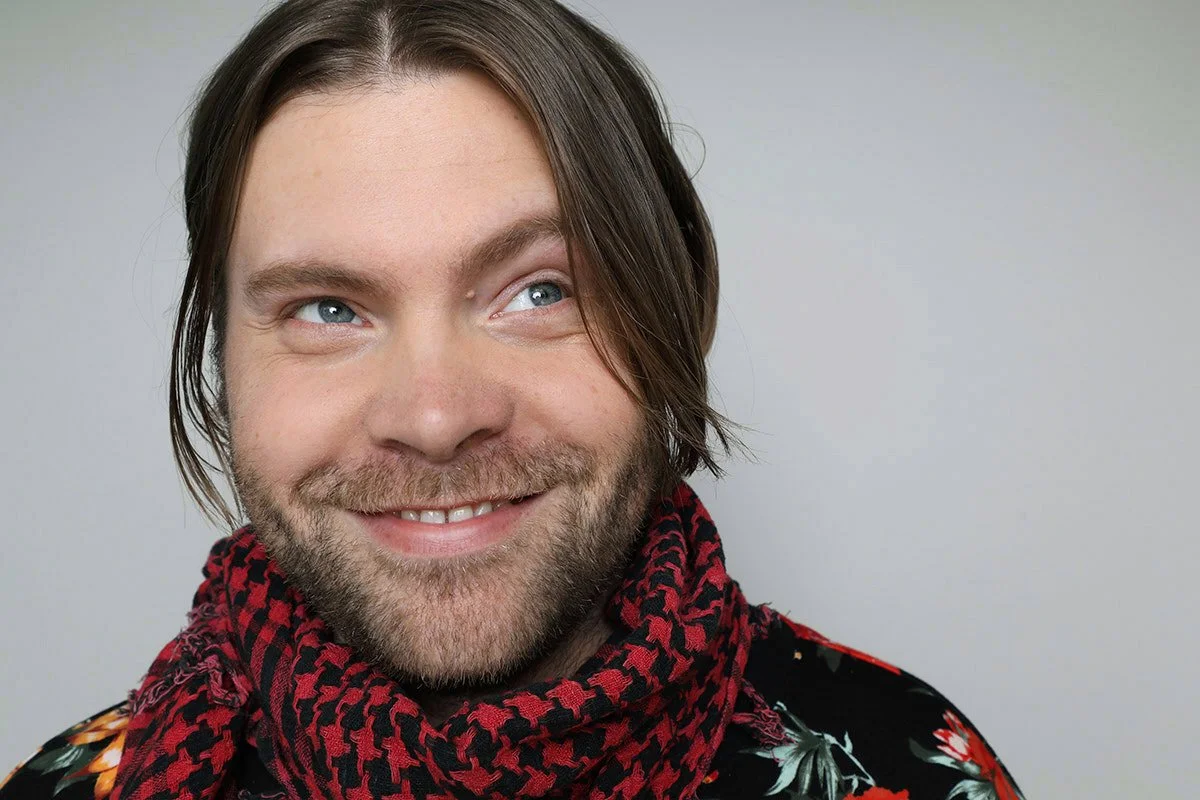 A smiling man with shoulder-length brown hair, a beard, and blue eyes, wearing a black shirt with floral patterns and a red and black checkered scarf.