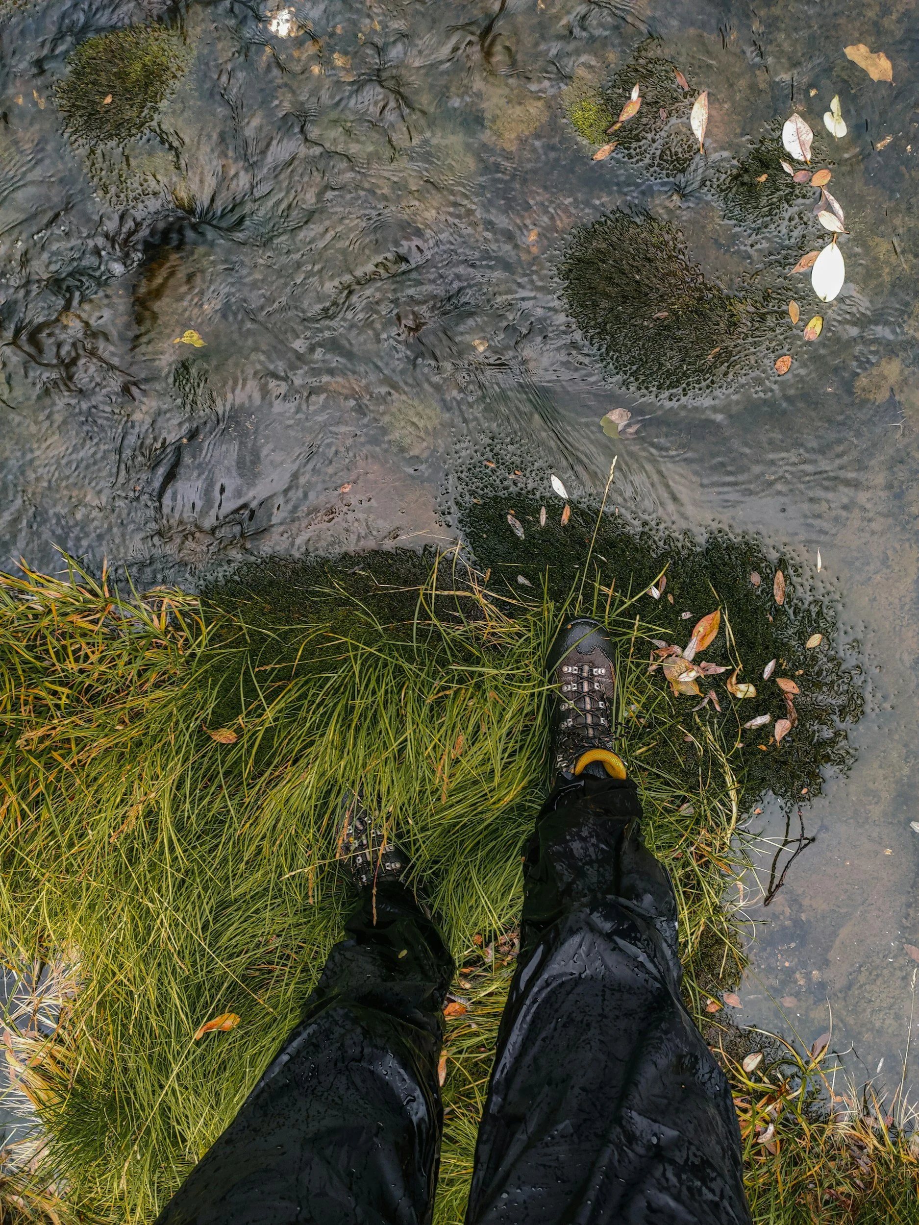 man's feet in hiking boots standing on grassy edge by running stream