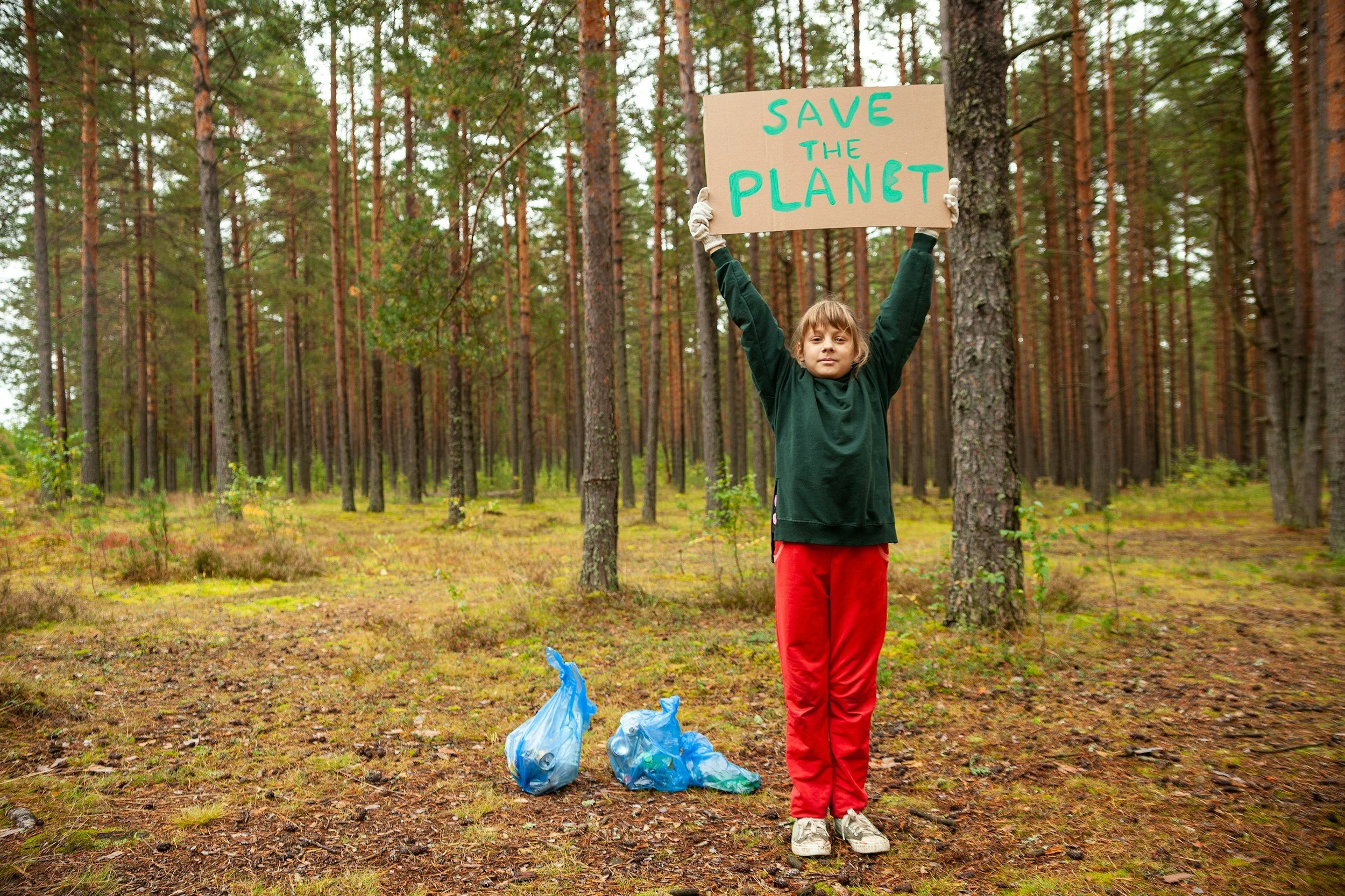 young girl in green sweatshirt and red sweatpants and sneakers holding up save the planet sign above her head while standing in forest with two blue garbage bags at her feet