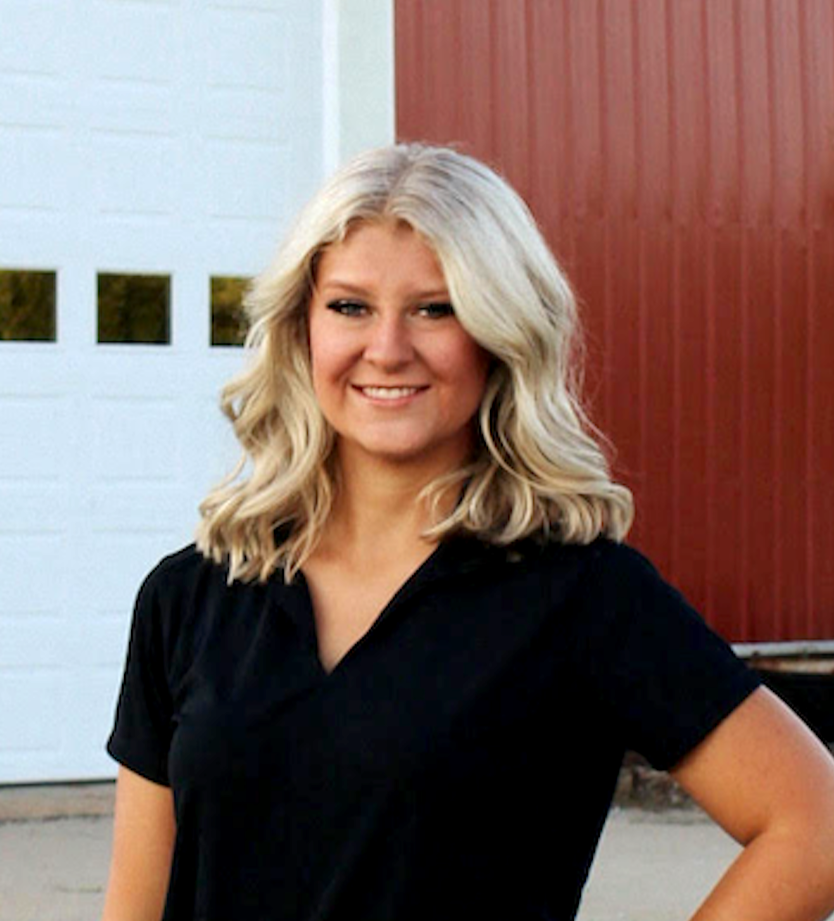 young blonde woman smiling in black v-neck shirt standing outside in front of red pole barn with white garage door and small square windows