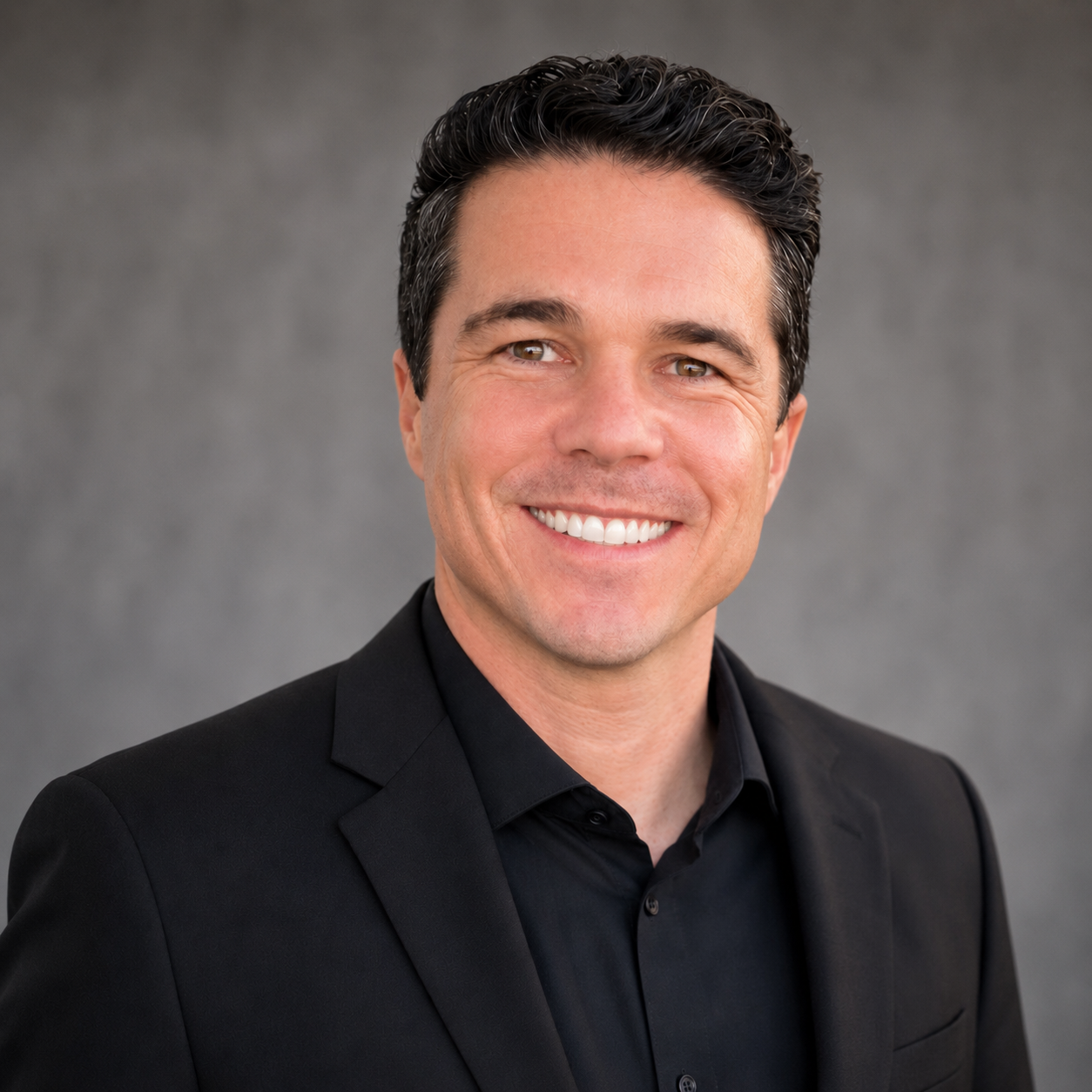 Headshot of a smiling man in a black suit against a gray background.