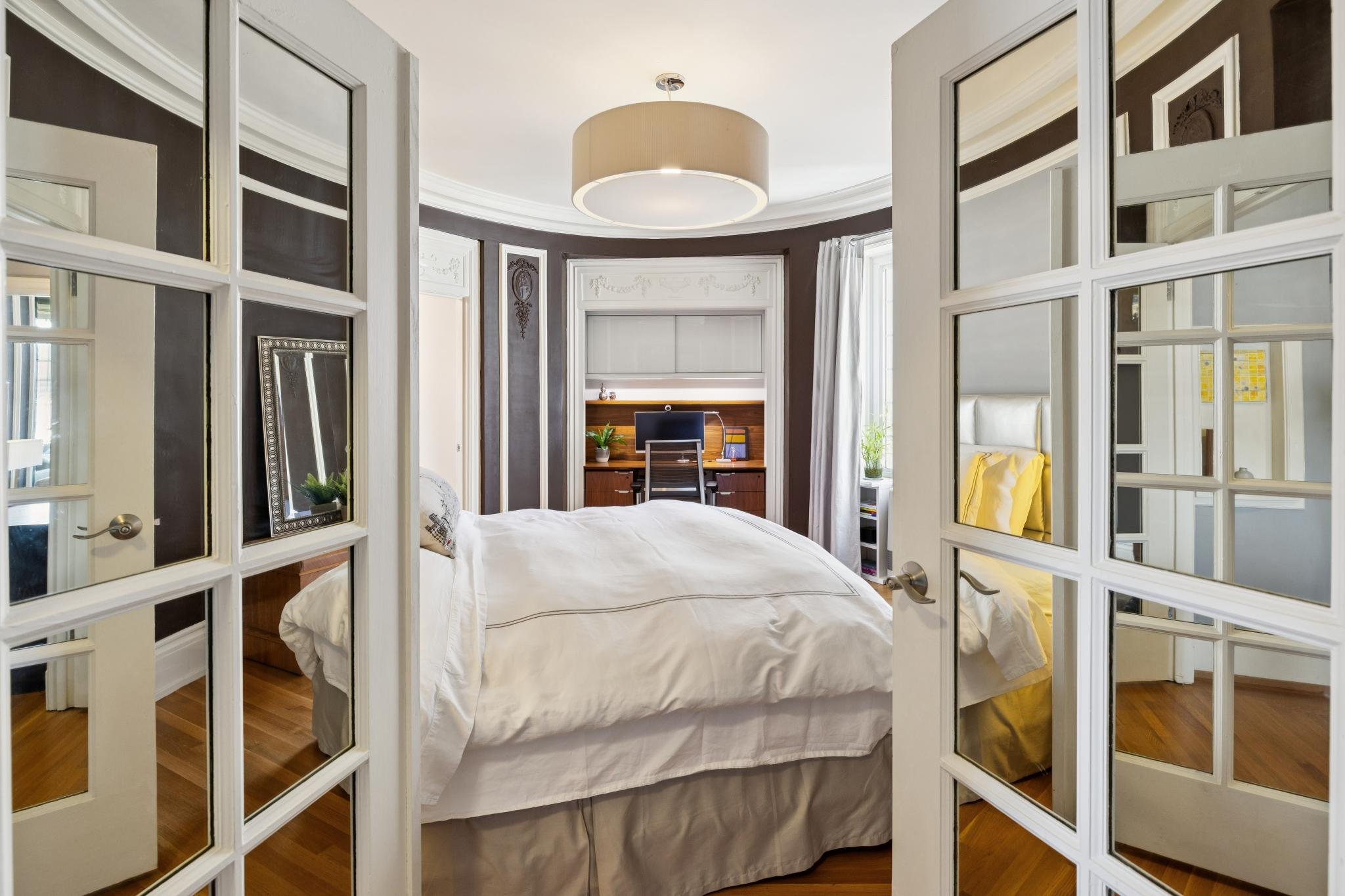 A bedroom seen through a glass-paneled folding door, featuring a bed with white bedding, a wooden desk with a computer, a window with white curtains, and decorative molding on the walls.