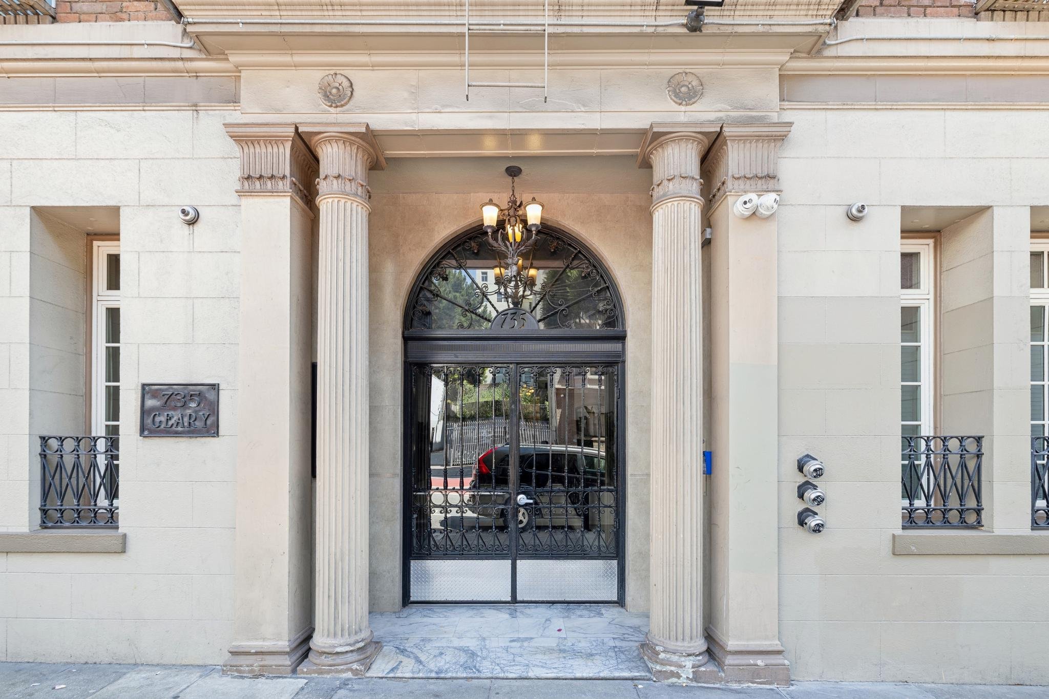 Facade of a beige building with two large columns framing a wrought iron gate and a doorway above bearing the number 735. Above the entrance hangs a chandelier with five lights, and small security cameras are mounted on the walls.