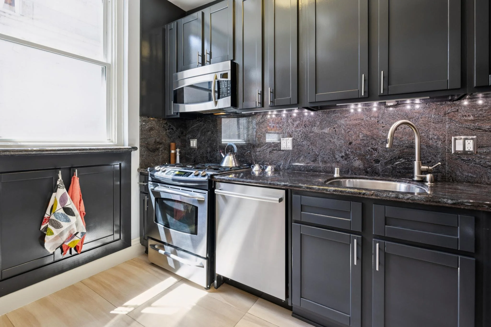 A modern kitchen with dark cabinets, a granite countertop, and stainless steel appliances including a microwave over the stove. A tea kettle is on the stove, and a colorful dish towel hangs from the oven handle. There are electrical outlets on the ba