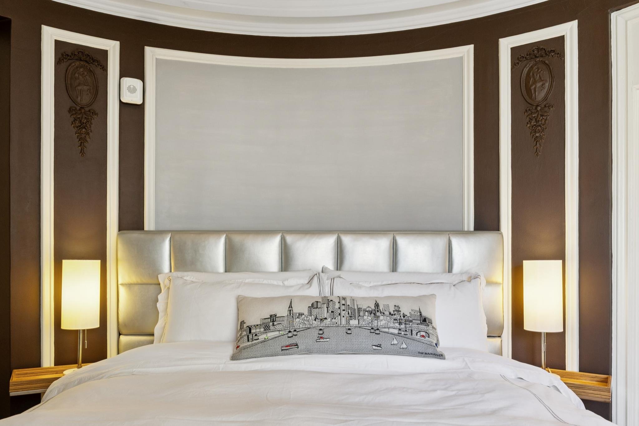 A modern bedroom with a large bed, white bedding, and a decorative pillow featuring a cityscape illustration. The headboard is padded and behind it is a brown wall with white trim and two decorative panels on either side. There are two tall lamps on 