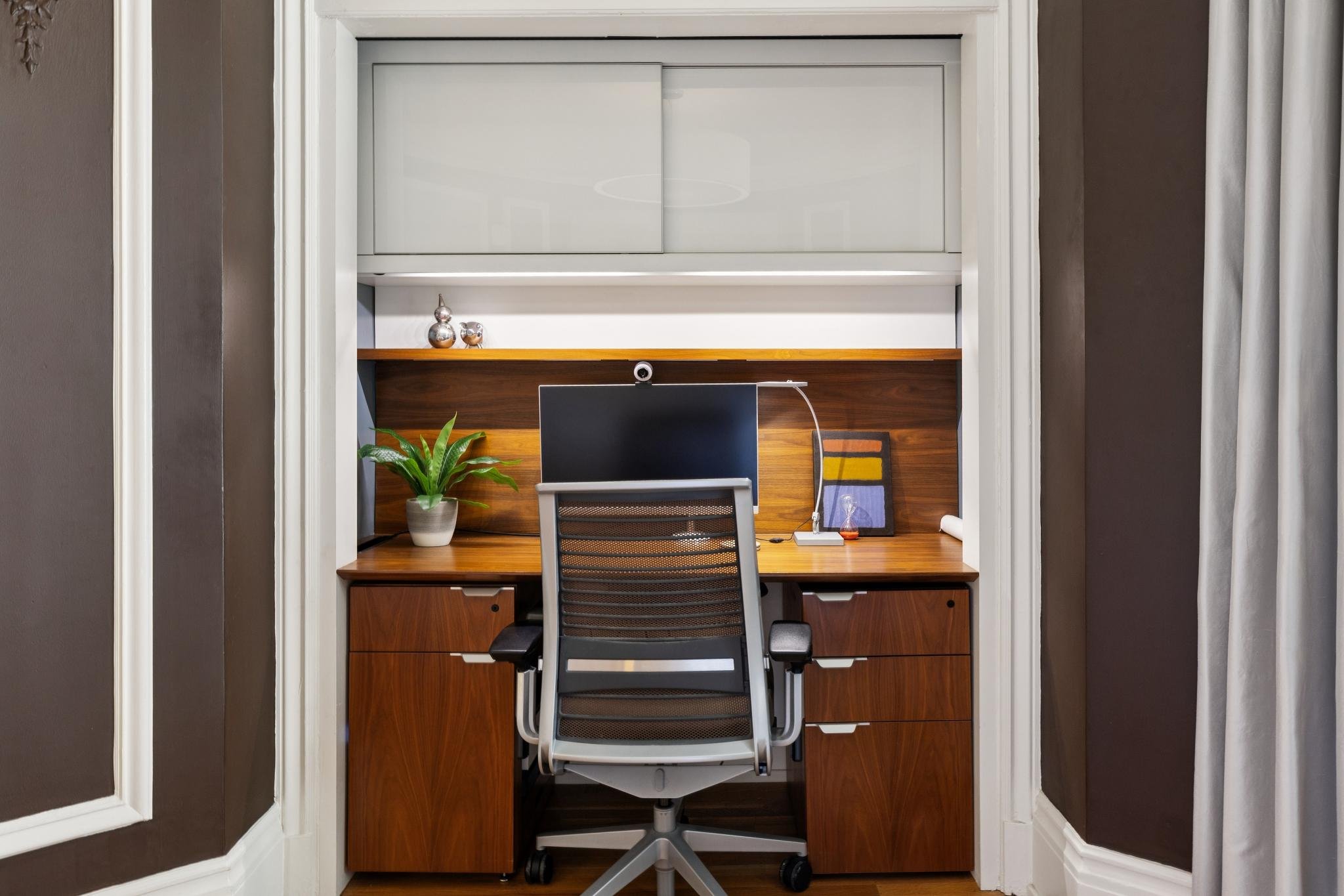 A home office setup viewed through a doorway. It features a wooden desk with a computer monitor, a potted plant, a webcam, and decorative items. There are white and wood-paneled wall features, and a gray chair is in front of the desk.