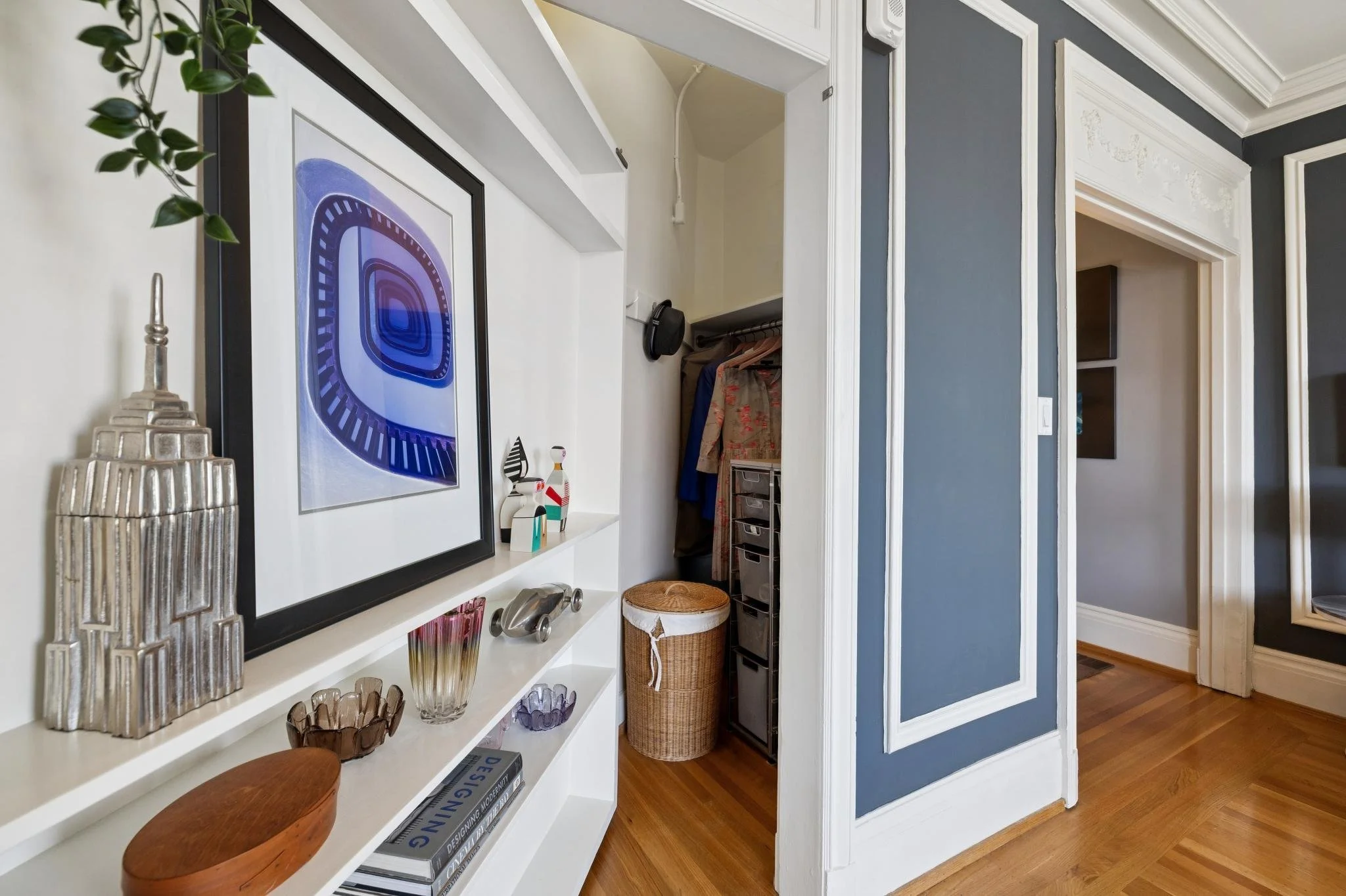 Interior view of a residential living area showing a white shelf with decorative items such as glassware and a framed artwork, overlapping a section with blue wall panels and a doorway leading to a walk-in closet with clothes hanging and plastic stor