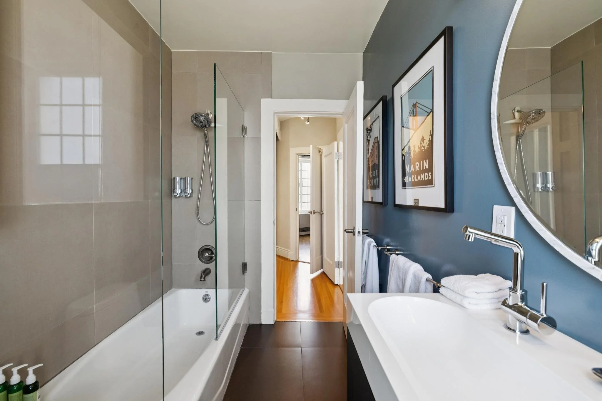 A modern bathroom with a walk-in shower with glass doors and a handheld showerhead, a white vanity with a round mirror, and artwork on a dark blue wall. The bathroom has hardwood floors and a doorway leading to a room with a window.