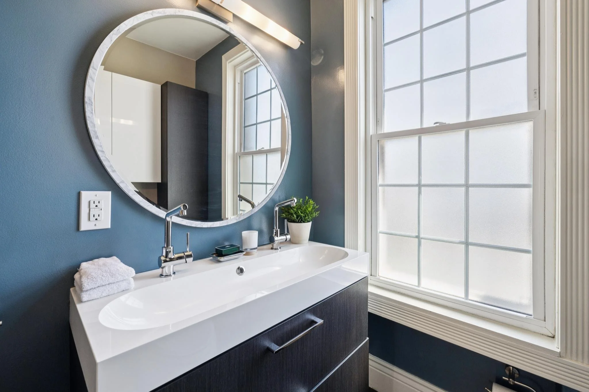 A modern bathroom vanity with a white sink, chrome faucets, a black cabinet, a round mirror, and a large window with a grid pattern letting in natural light.