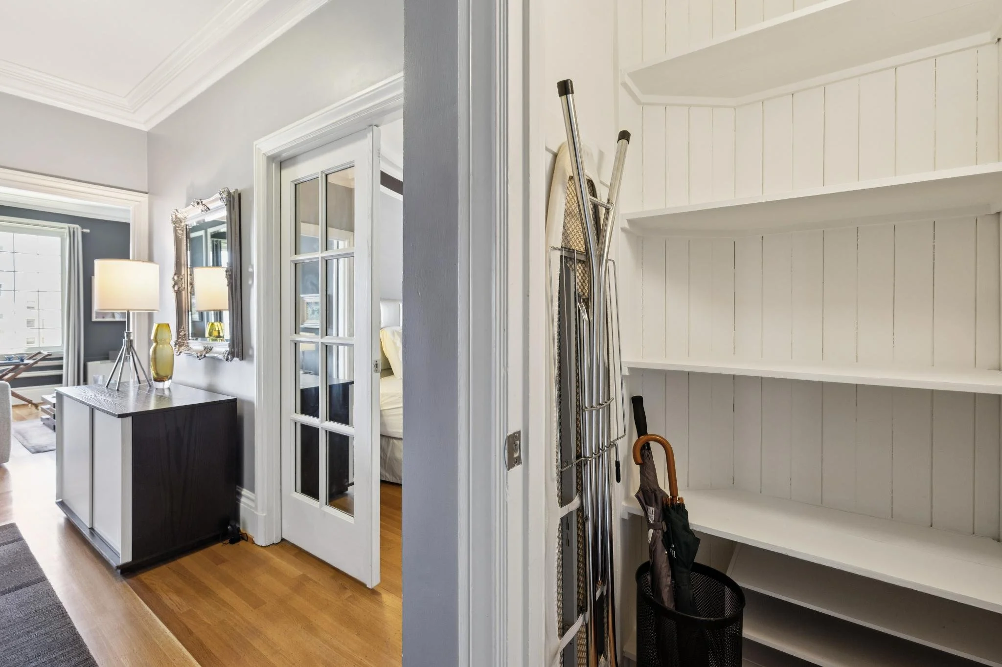 A corner of a room with white shelving, on which are stored umbrellas and a backpack, with a laundry room visible through an open door at the center.