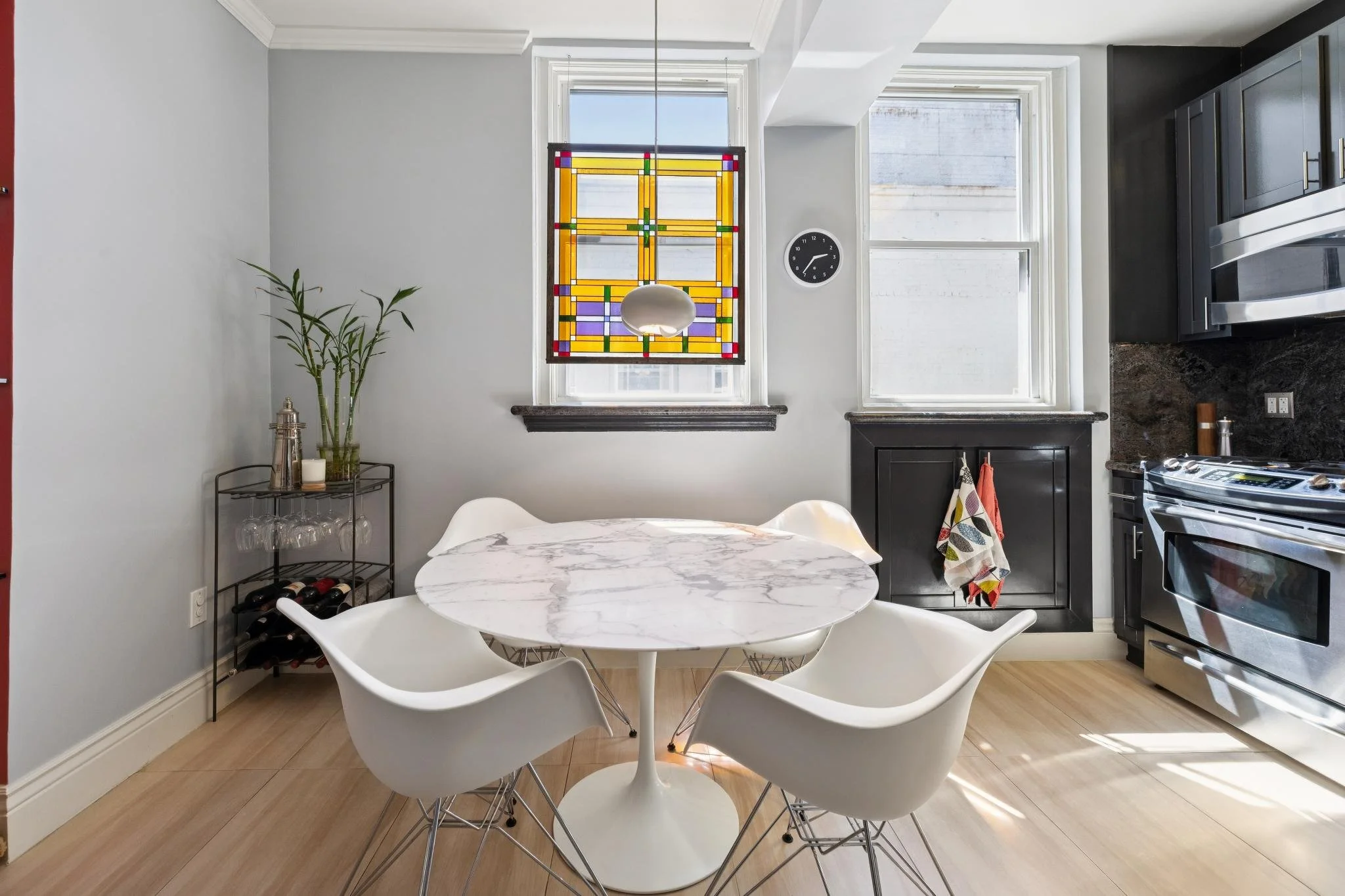 Modern kitchen with a marble dining table surrounded by white chairs, black cabinetry, a wine rack, colorful stained glass window, and two large windows with sunlight.