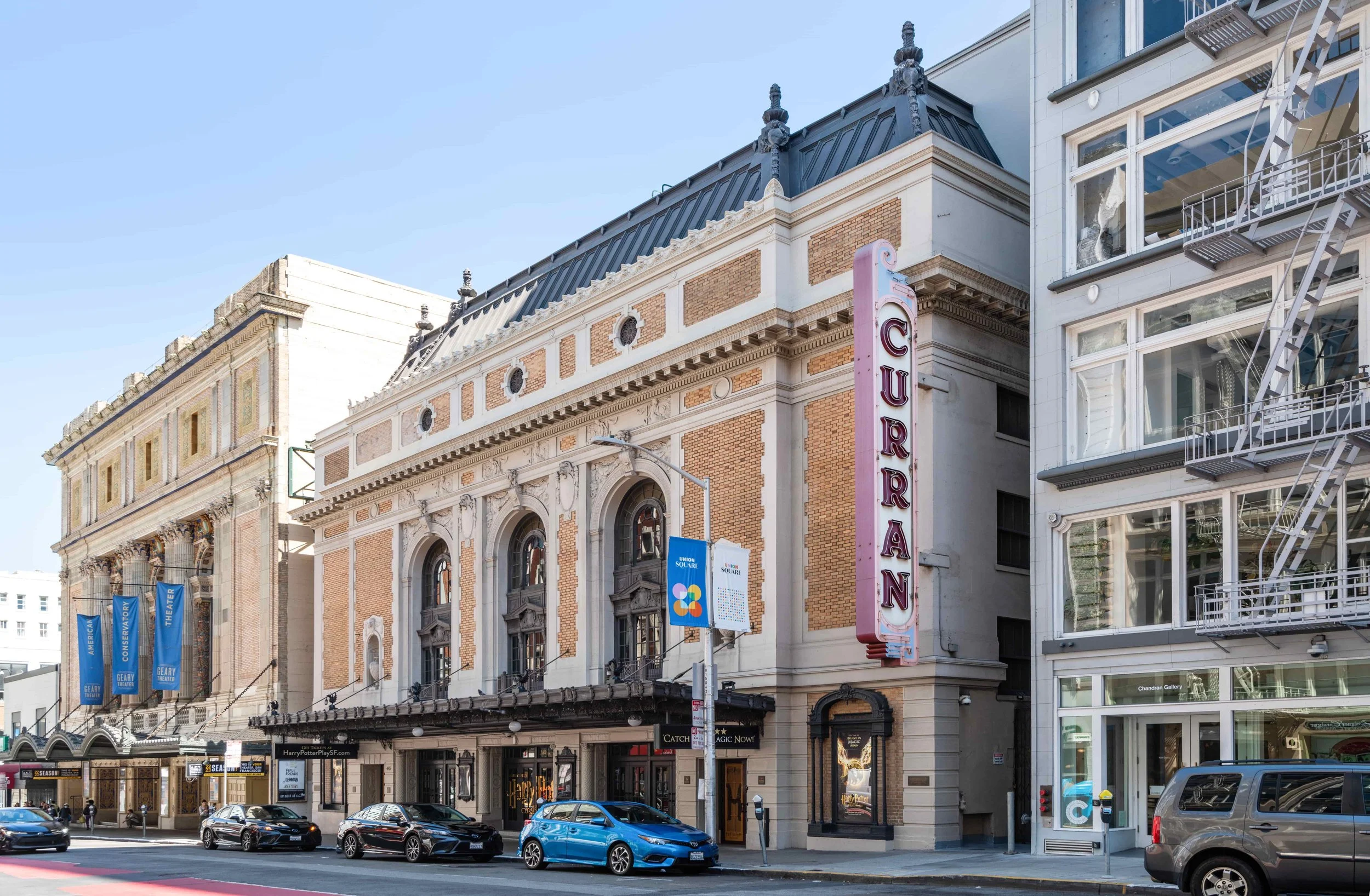 Street view of a historic theater building with a marquee, colorful banners, and cars parked in front. The building has vintage architecture with ornate details and arched windows.