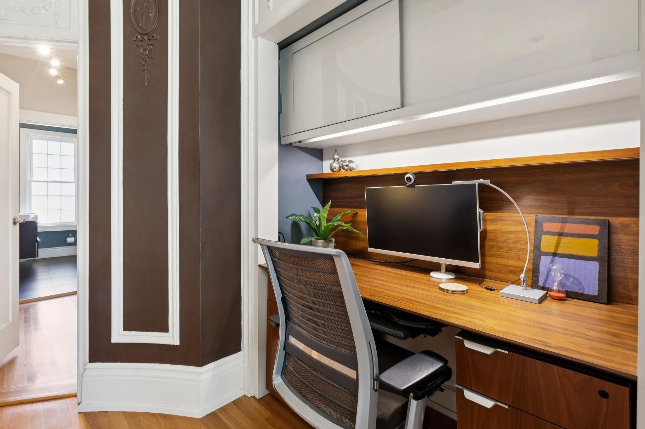 Home office with a wooden desk, computer monitor, chair, and decorative objects, near a wall with wood paneling and a shelf, with a window and hallway visible in the background.