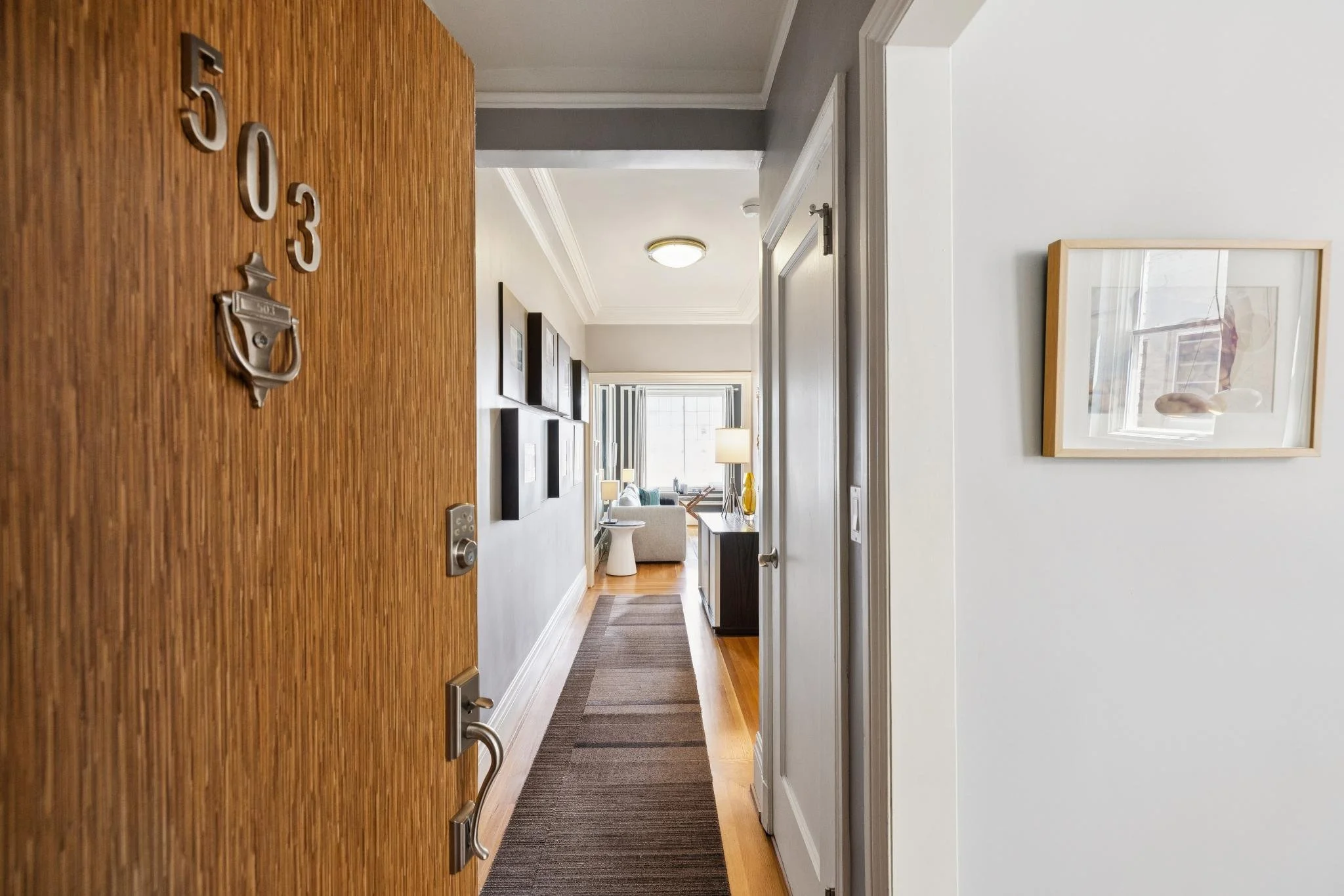 Open wooden front door with house number 503 mounted on it, leading into a hallway with wooden flooring, a striped rug, picture frames on the wall, a table lamp, and a view into a living room with large window, couches, and artwork.