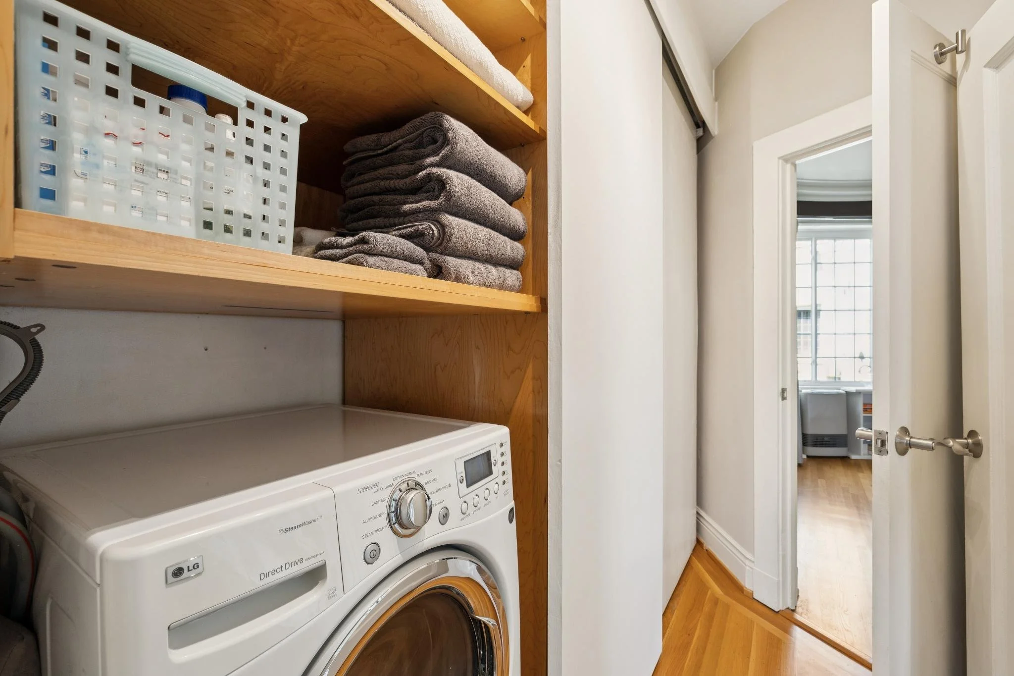 A laundry area with a front-loading LG washing machine and a shelf with folded towels, in a hallway leading to a room with a window.