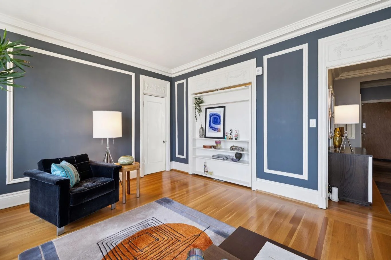 Living room with blue walls, hardwood floor, black velvet sofa with striped pillow, white built-in shelves with art and decor, white door, gray carpet with geometric design, side table with lamp and decorative objects, partial view of hallway with la