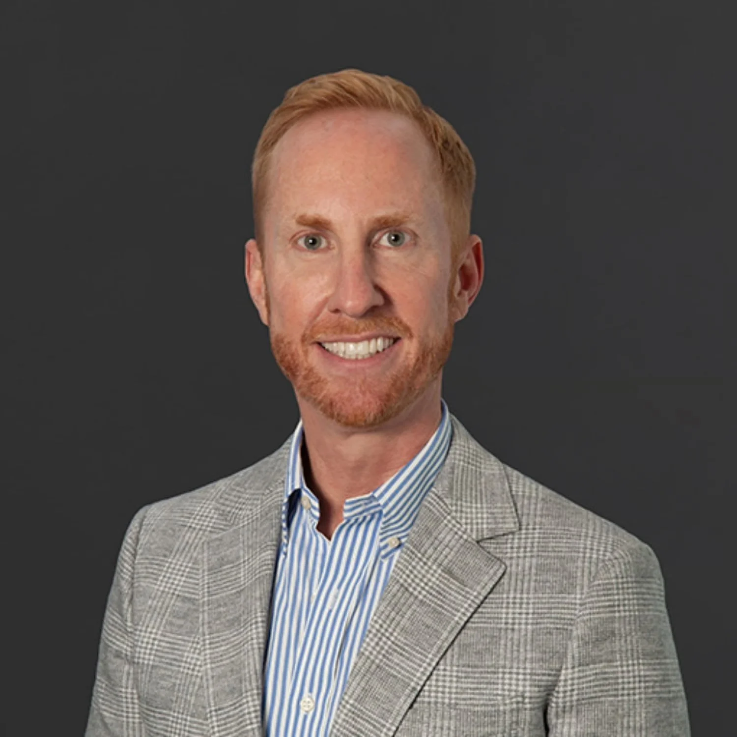 A portrait of a man with short red hair and a beard, wearing a gray checkered blazer and a blue striped shirt, smiling against a dark gray background.