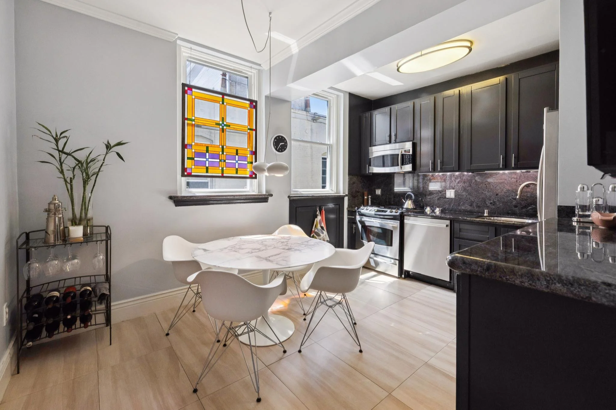 Kitchen with white ceiling and walls, black cabinets, stainless steel appliances, marble dining table with chairs, and windows with stained glass and natural light.