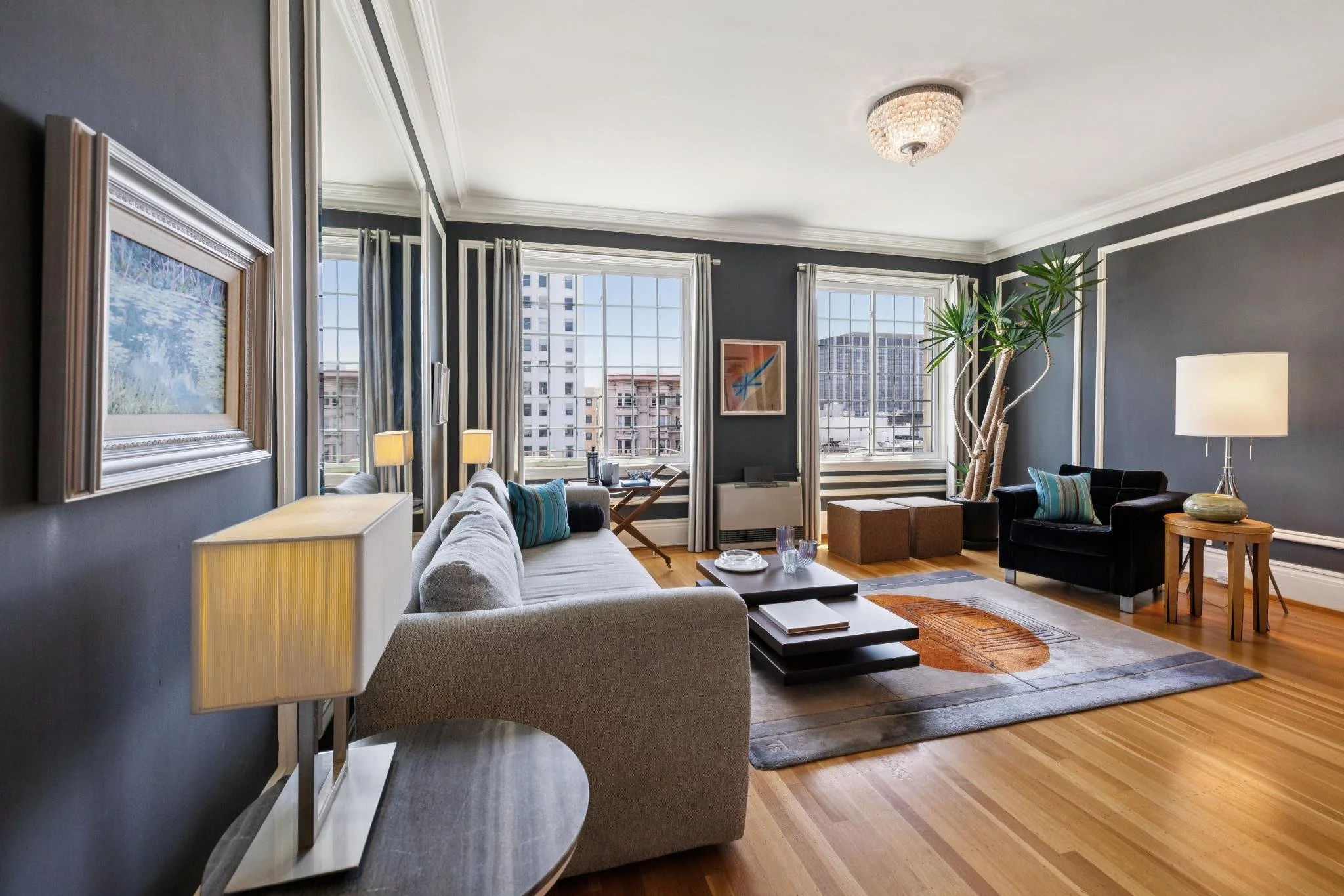 Living room with dark gray walls, large windows with white frames and curtains, light gray sofa, black armchair, wooden coffee table, colorful abstract artwork, plants, lamps, and hardwood flooring.