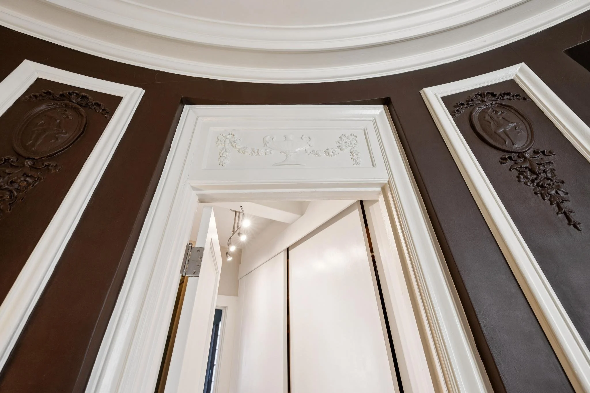 Interior view of a doorway with ornate dark brown and white molding, leading into a well-lit room with ceiling lights visible.