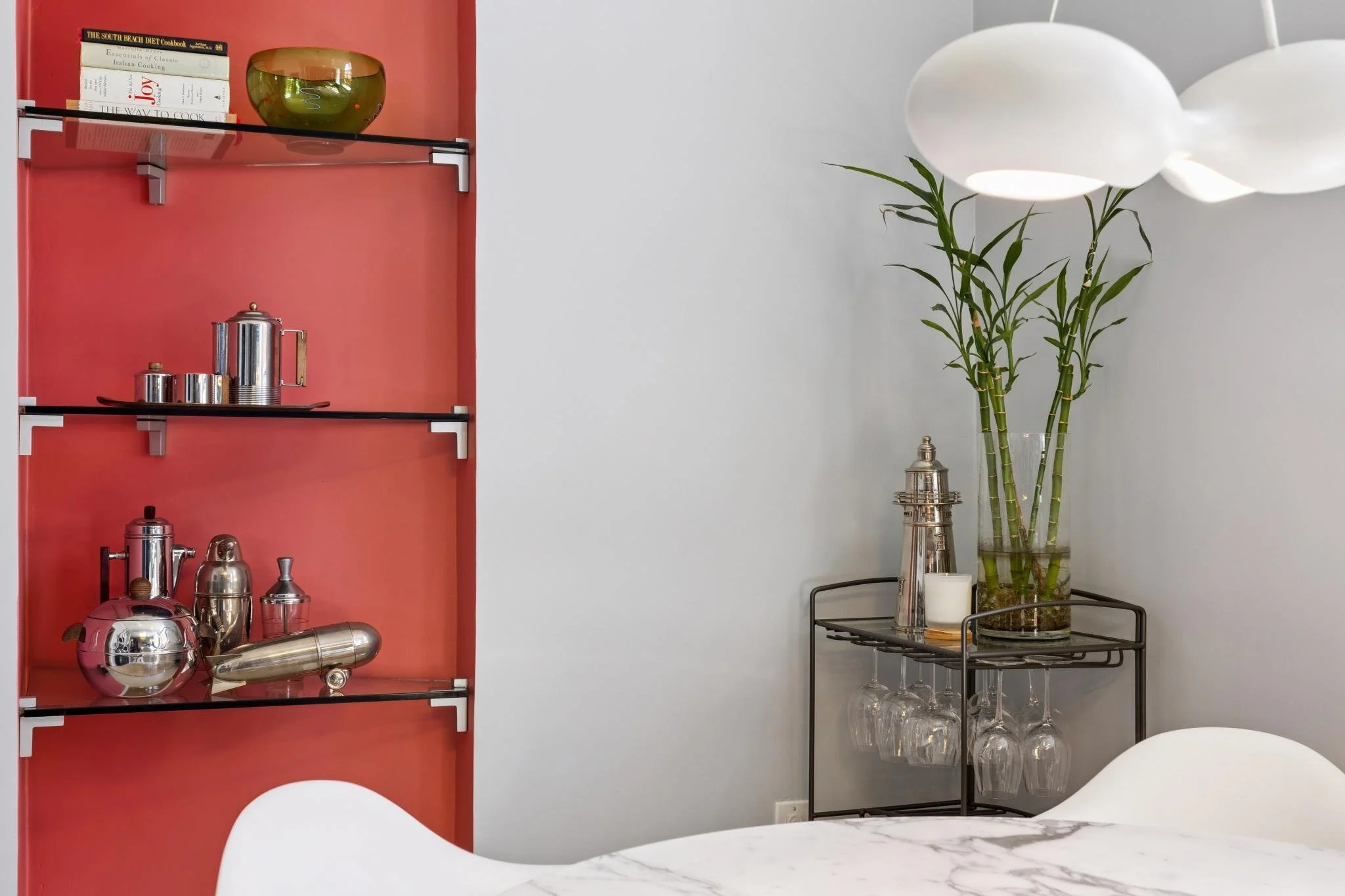 A red wall-mounted shelf with books, a green glass bowl, and silver tea or coffee pots. Next to it is a tall glass vase with bamboo stalks and white orchids, set on a black metal side table with glassware and candles.