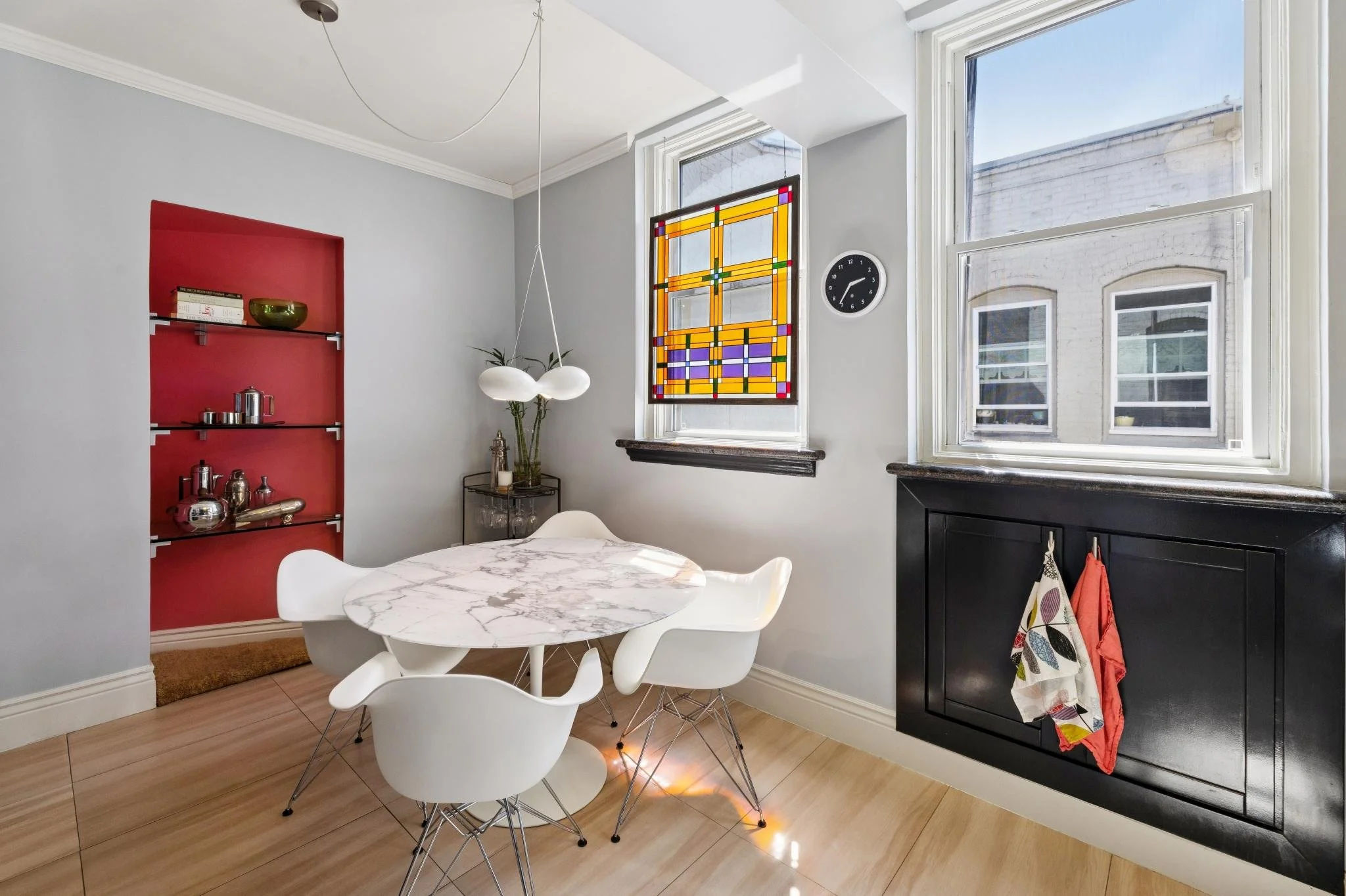 A dining area with a marble-top table and white modern chairs, a wall with a red built-in shelf displaying decorative items, a large window with a stained glass hanging, and a black cabinet with utensils and colorful towels hanging.