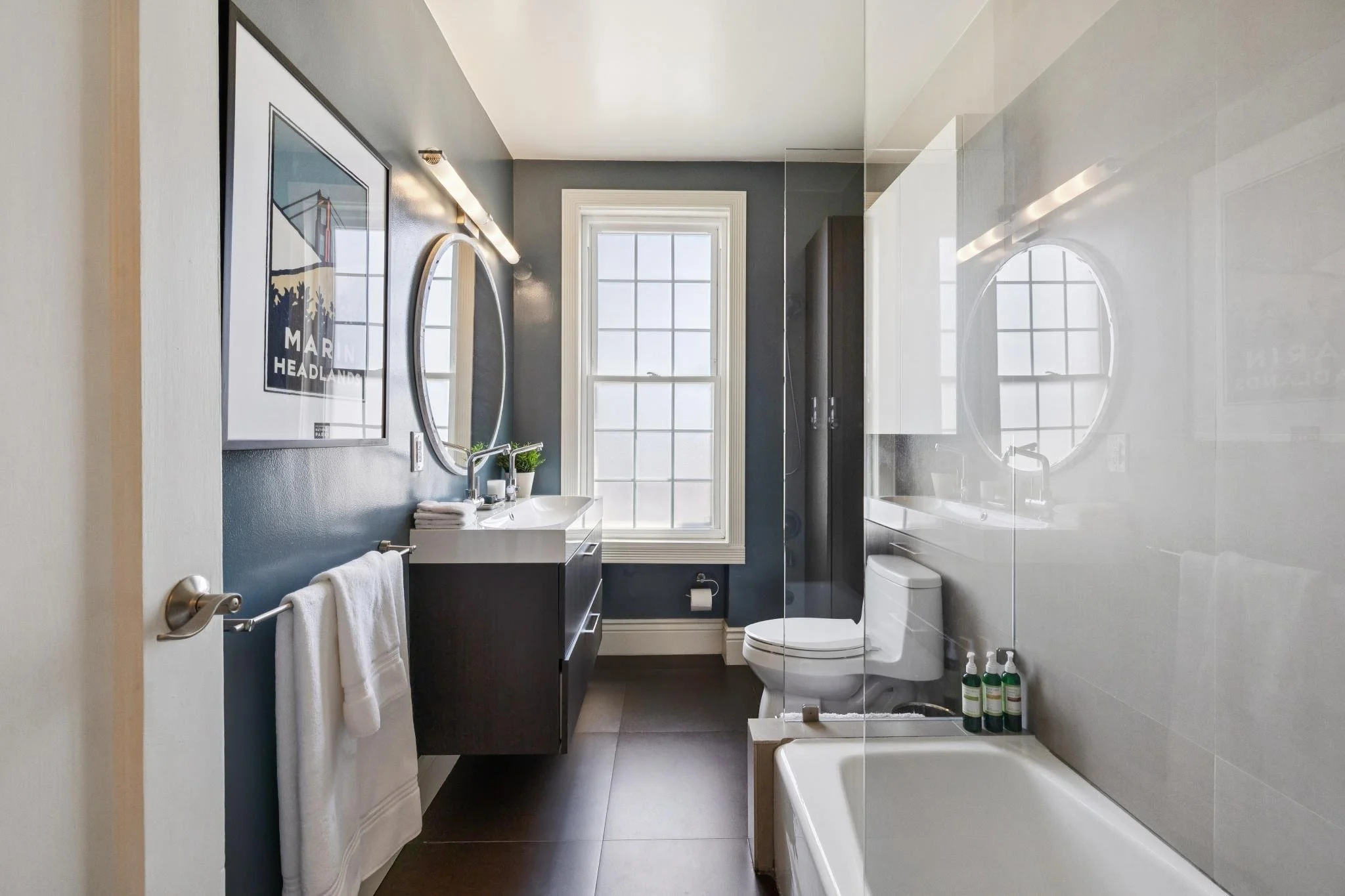 Modern bathroom with dark vanity, white sink, oval mirror, window, bathtub, and toiletries.