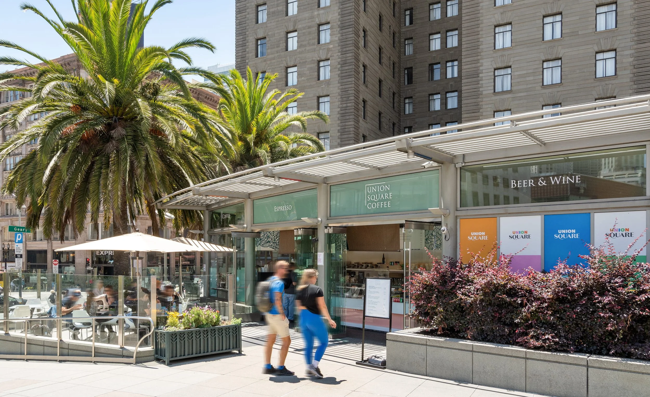 People walking past a coffee shop named 'Union Square Coffee' with palm trees and colorful flowering bushes in front, and tall buildings in the background.