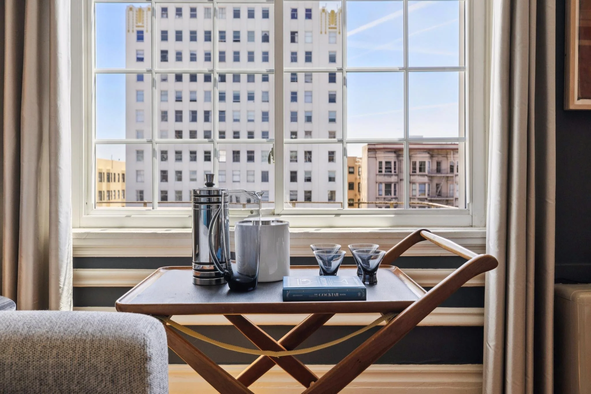 A view of city buildings through a large window from inside a room, with a wooden table set with a coffee pot, coffee cups, and a book titled 'The Cocktail Book.'