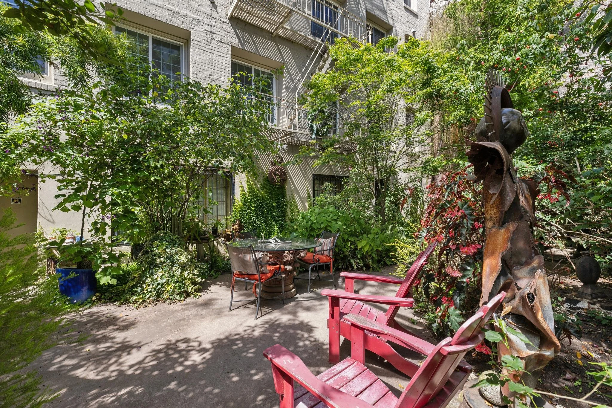 A cozy outdoor patio area surrounded by lush green foliage and trees, with various chairs and a glass table, and a metal sculpture of a face wearing a hat.