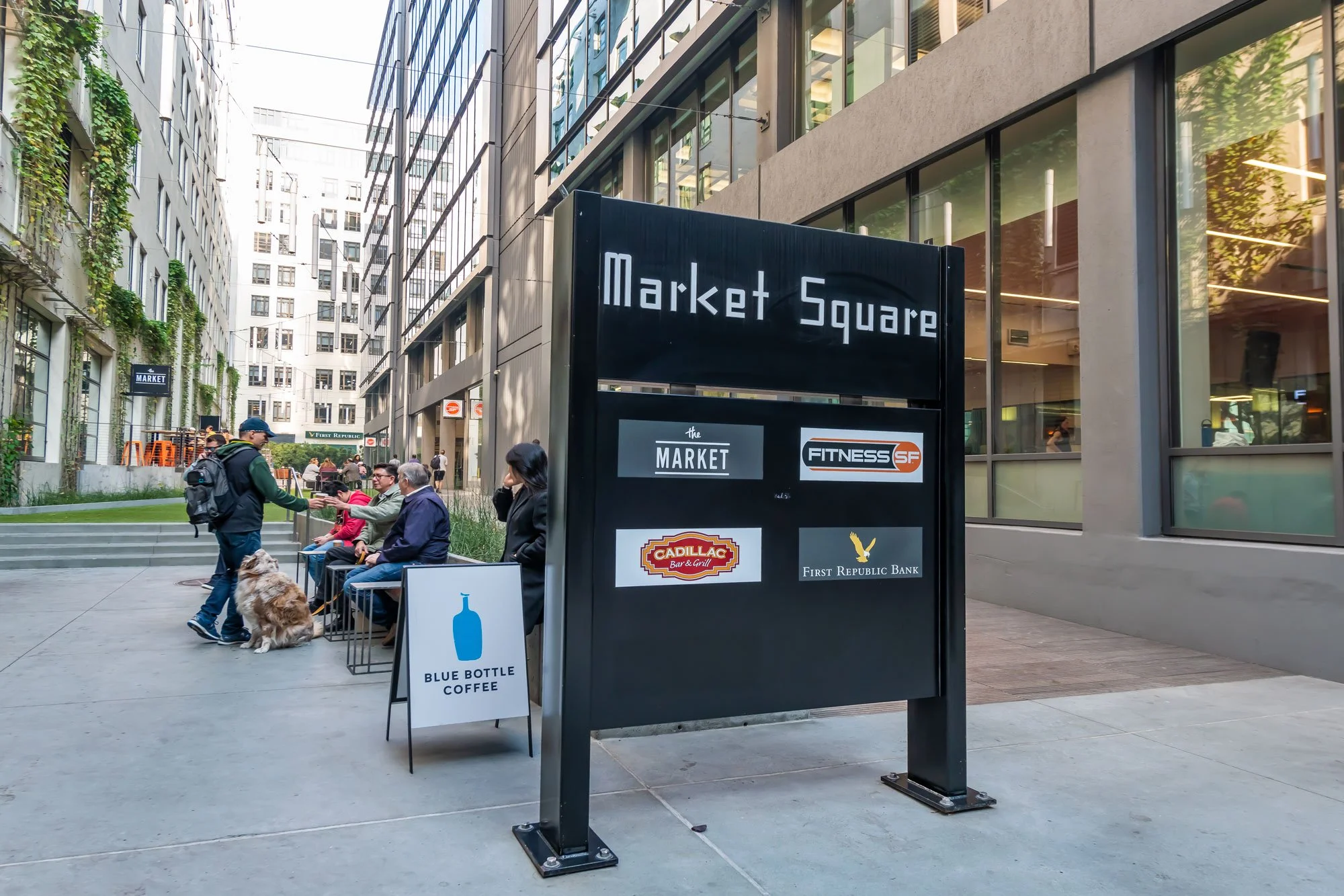 Outdoor seating area in a city with people, a dog, and a large sign for Market Square listing businesses such as The Market, Fitness SF, Cadillac Bar & Grill, and First Republic Bank. The area is surrounded by modern buildings with glass windows.