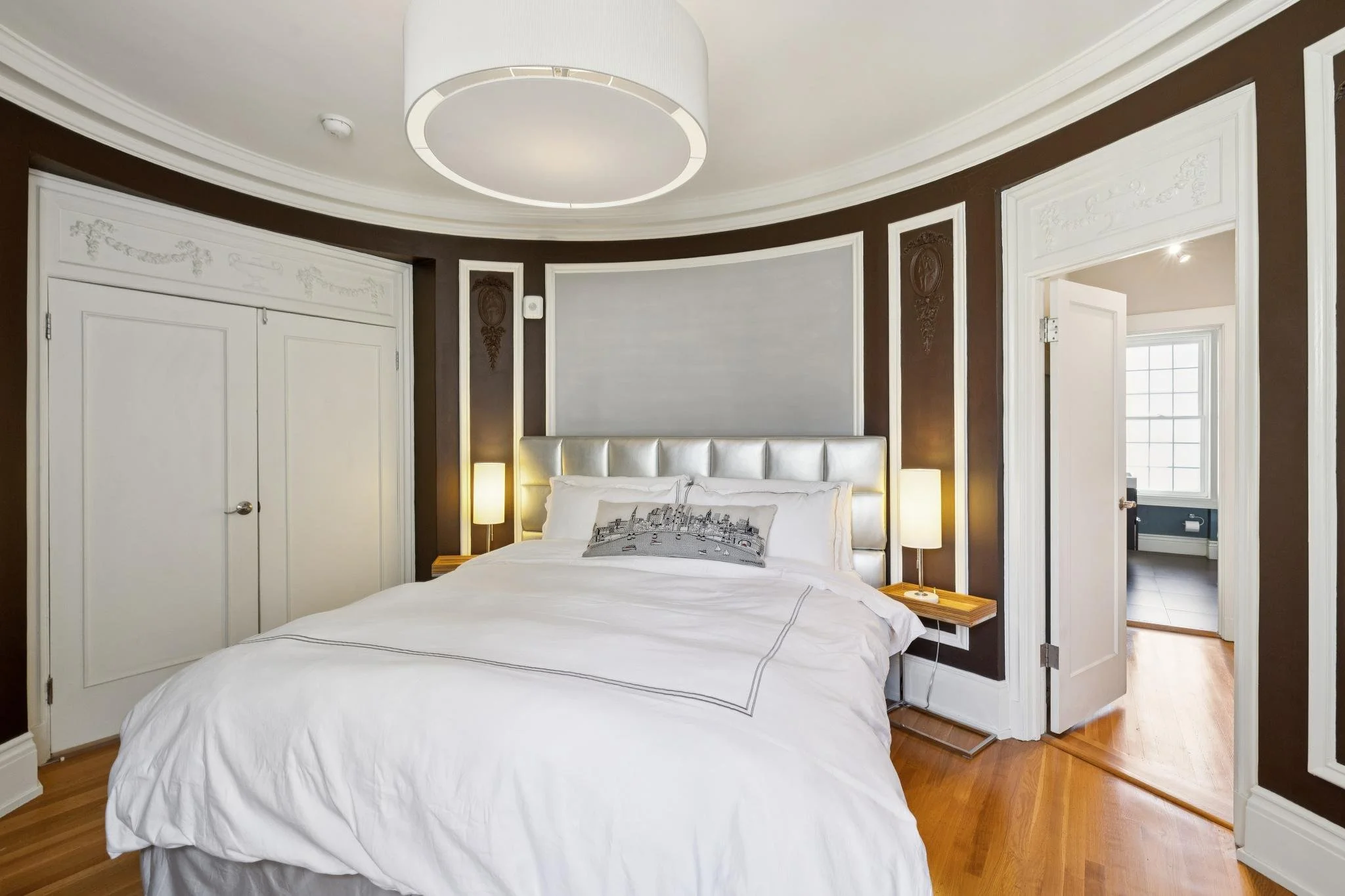 A bedroom with a large bed with white bedding and a decorative pillow featuring cityscape art, two bedside tables with lamps, a mirrored closet door, a dark accent wall with decorative medallions, and a doorway leading to another room with a window.