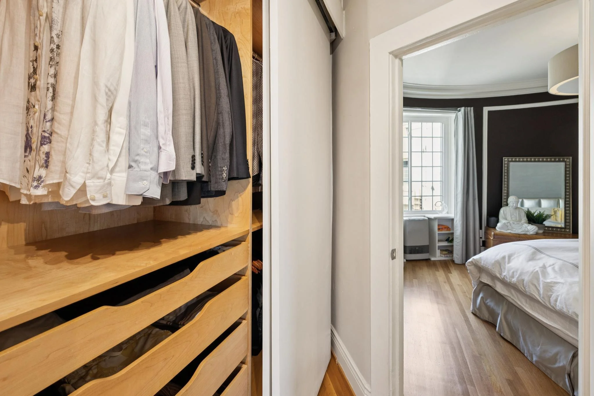 A wardrobe with neatly hanging shirts and jackets in front of a mirror reflecting a bedroom with a white bed, large window, and black wall.
