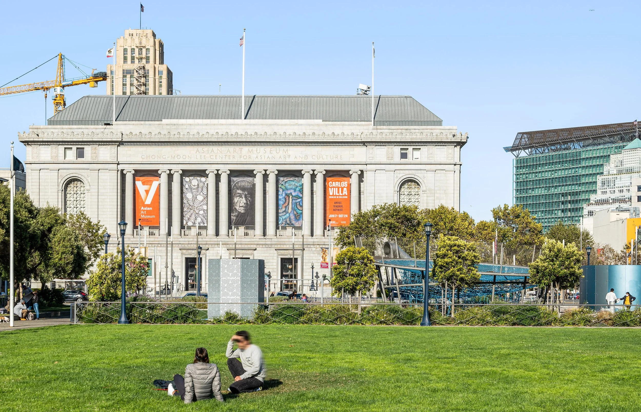 People sitting and talking on a green park lawn in front of a large building with columns and banners, with city skyscrapers and a construction crane in the background.
