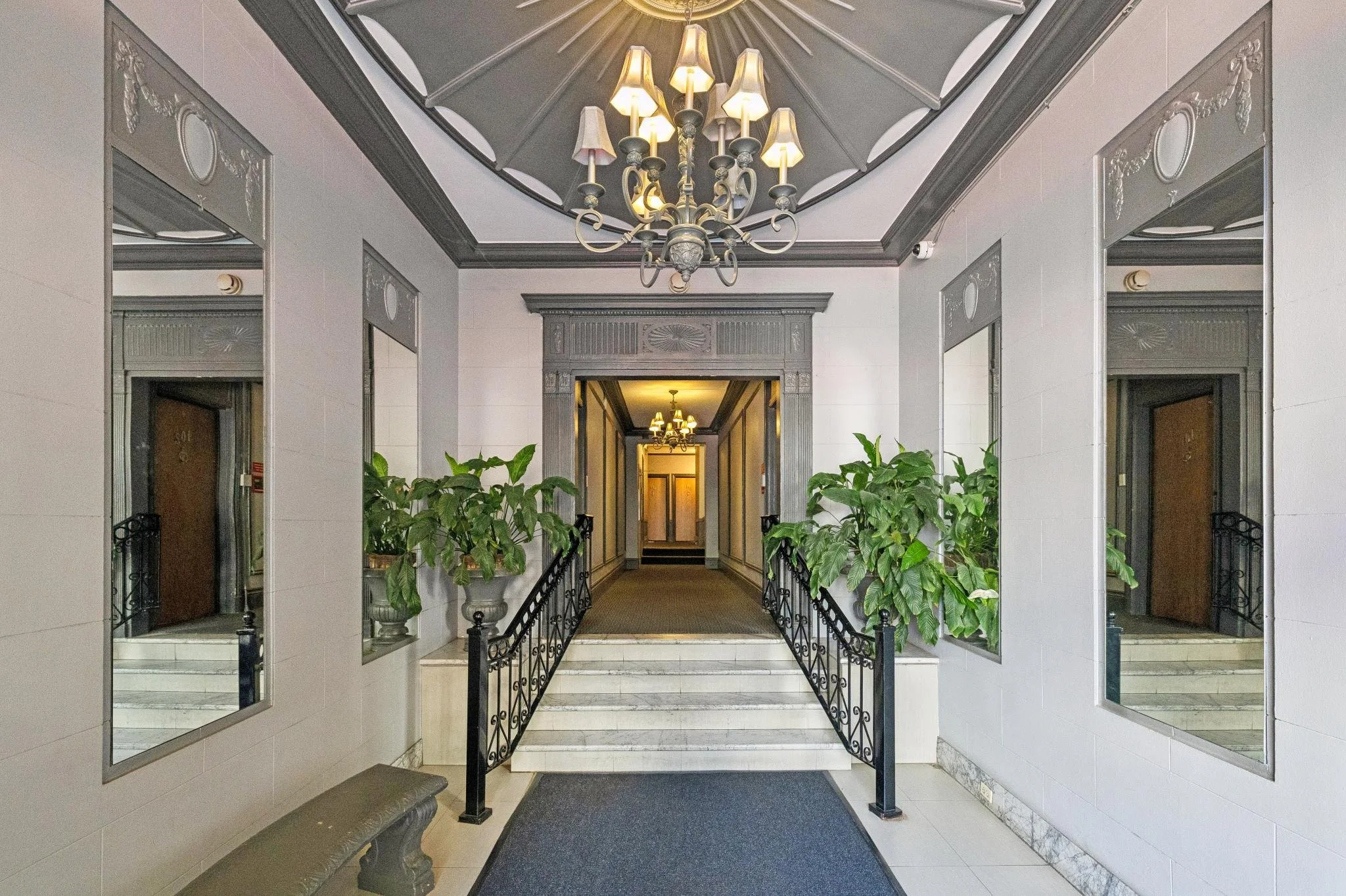 Indoor hallway featuring a chandelier with multiple lampshades hanging from the ceiling, surrounded by framed mirrors and potted plants along the railing, with a doorway at the end.
