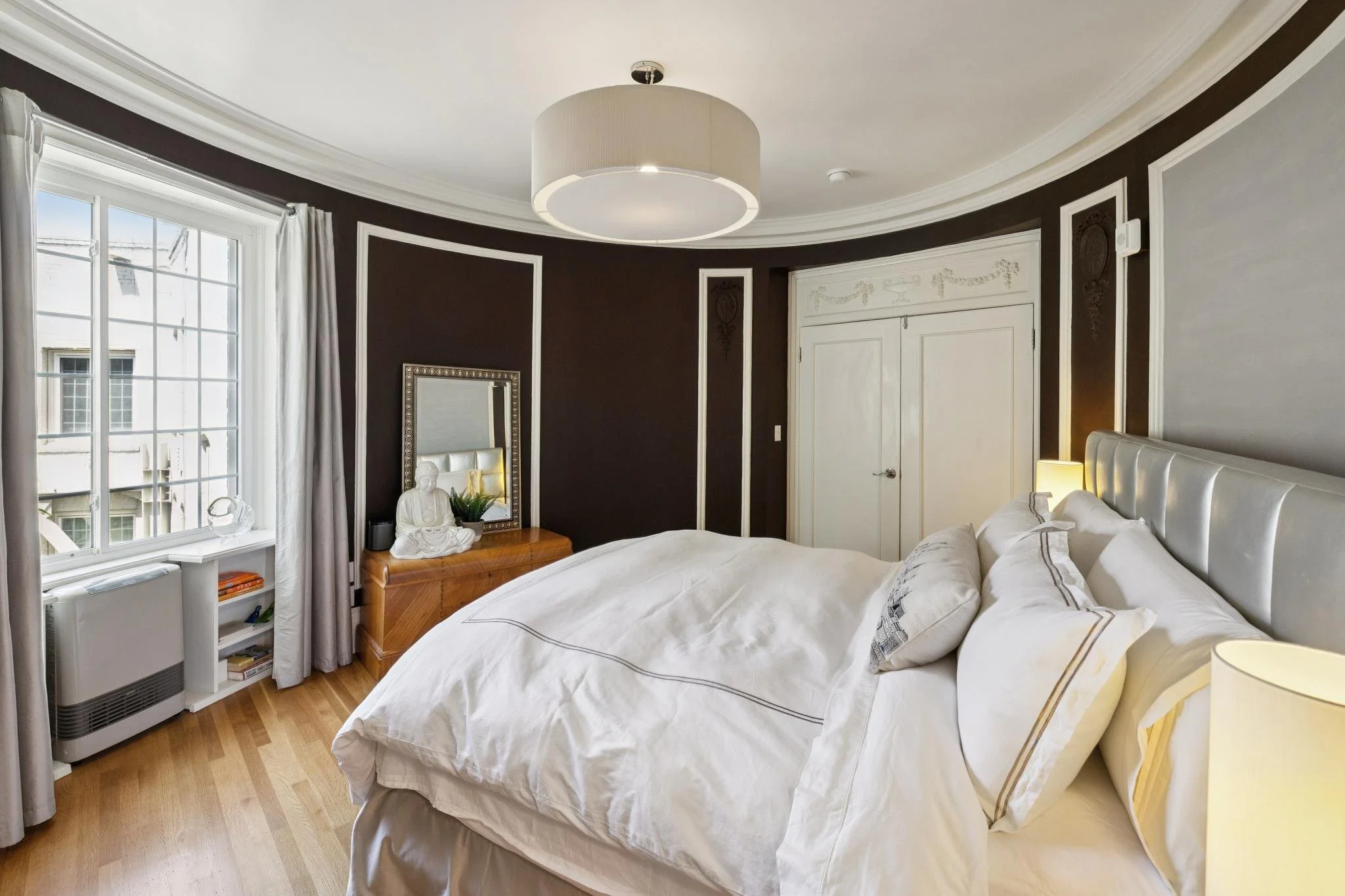 A bedroom with a large bed covered in white linens, a white padded headboard, a wooden side table with a Buddha statue, a mirror, a lamp, large windows with white curtains, and dark walls with white trim.