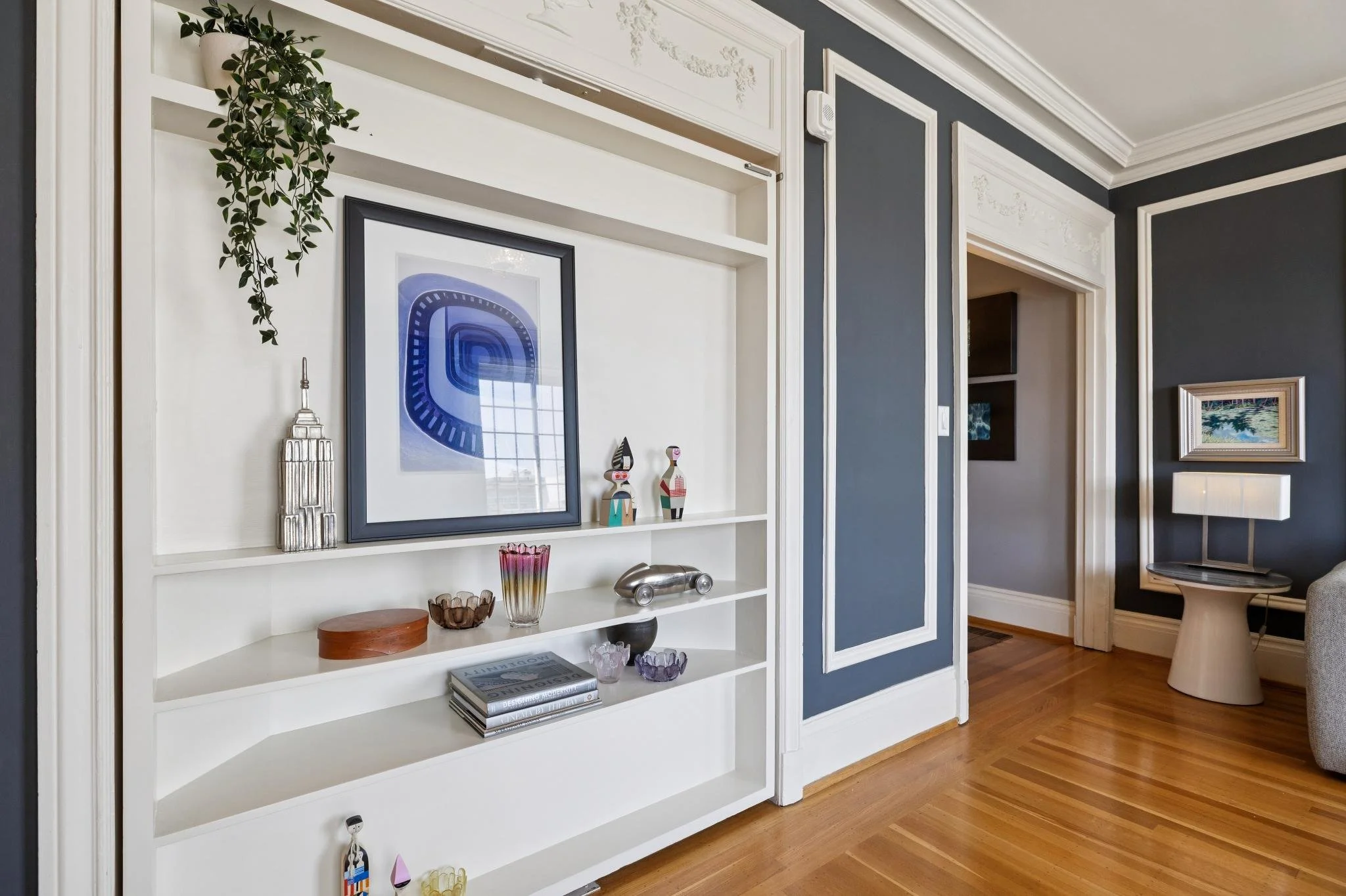 Interior view of a living room with a white built-in shelf unit decorated with framed artwork, small sculptures, vases, and books. There is a dark gray wall with detailed molding accents, hardwood floors, and additional artwork hanging on the wall.