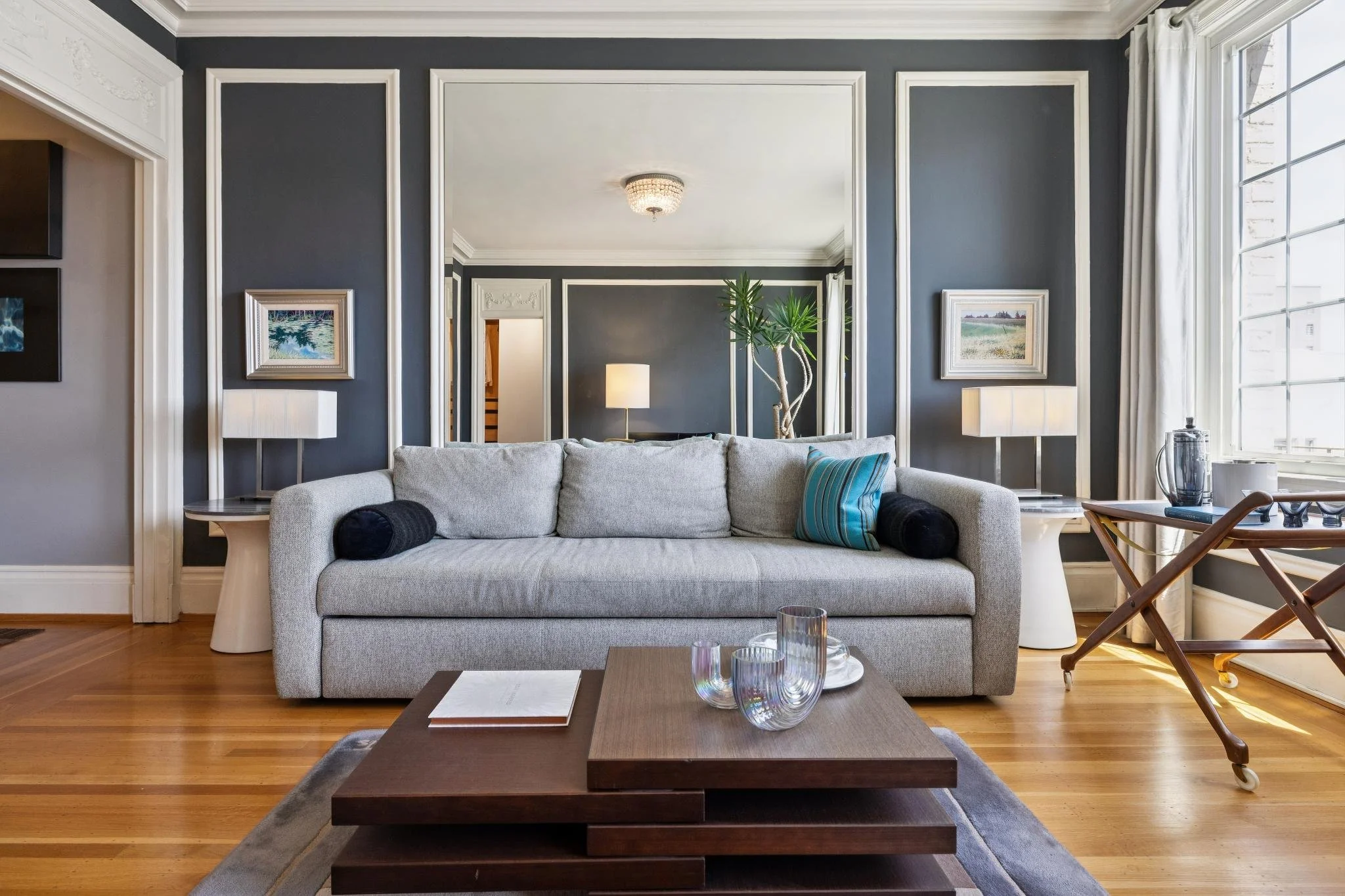 Living room with a white sofa, wooden coffee table, two side tables with lamps, art on dark gray walls, a plant, and a large window with curtains.