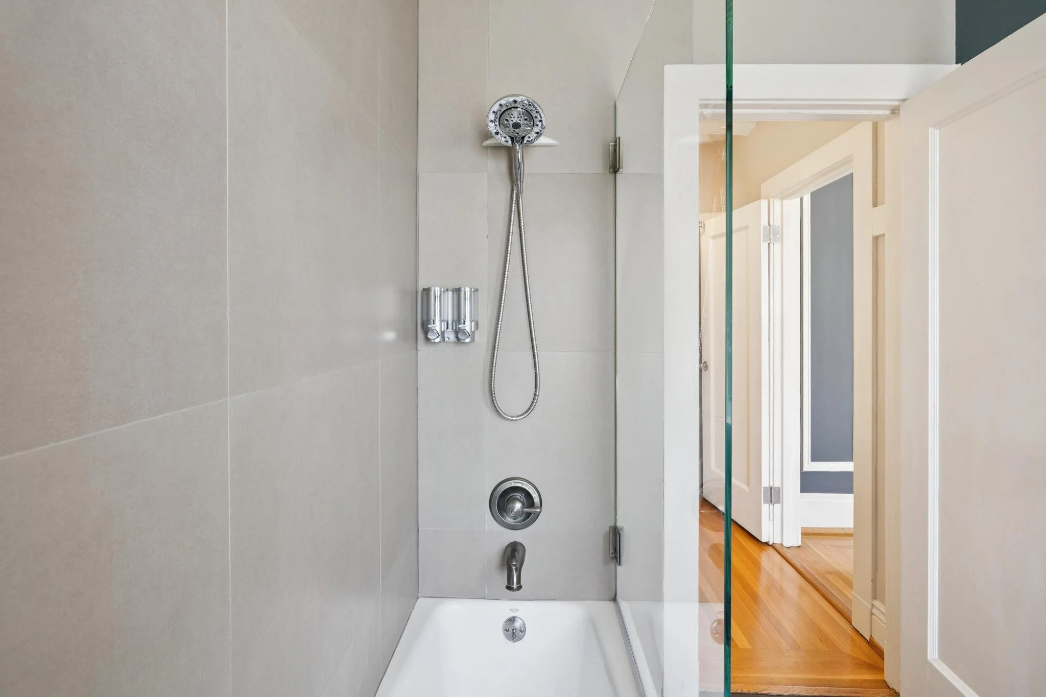 A modern shower area with beige tiles, a handheld showerhead, a metal drain, and a glass partition, with a view into a room with a wooden floor and a door.