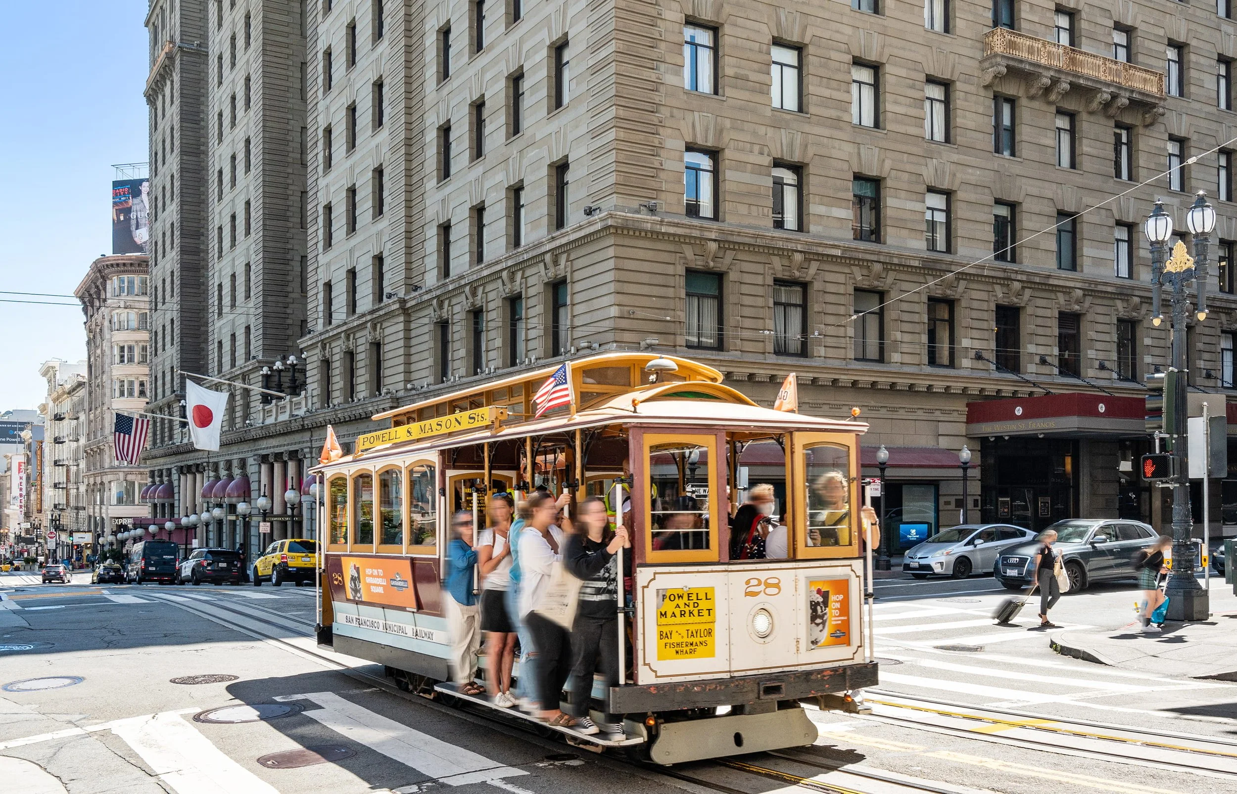 A cable car with passengers rides through a busy city intersection in San Francisco, California, with historic buildings and street signs visible.