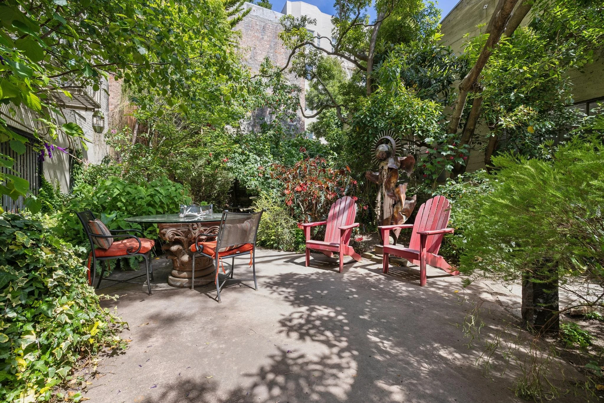 A lush, green backyard patio with trees and plants surrounding a concrete surface. There are outdoor chairs including red Adirondack chairs and black metal chairs around a stone table. Shadows from the trees cast patterns on the ground. A decorative 