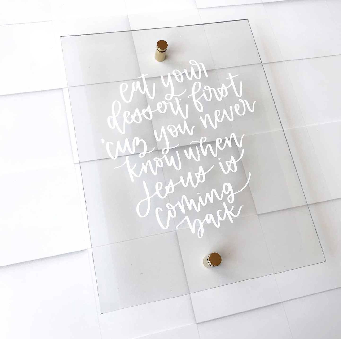 A clear acrylic sign with handwritten white text that reads, 'eat your dessert first, in case you never know when it is coming back.' The sign is mounted on a white tiled wall with small round standoff mounts.